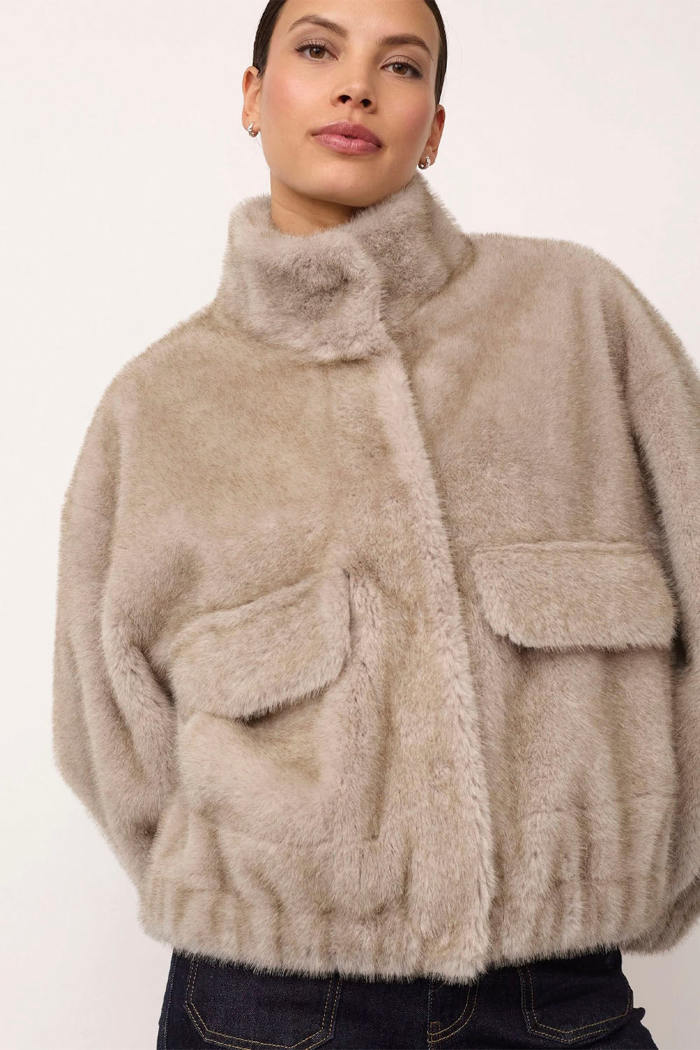 A woman wearing a beige fuzzy faux fur jacket with a high collar and flap pockets, paired with dark blue jeans, against a plain off-white background.