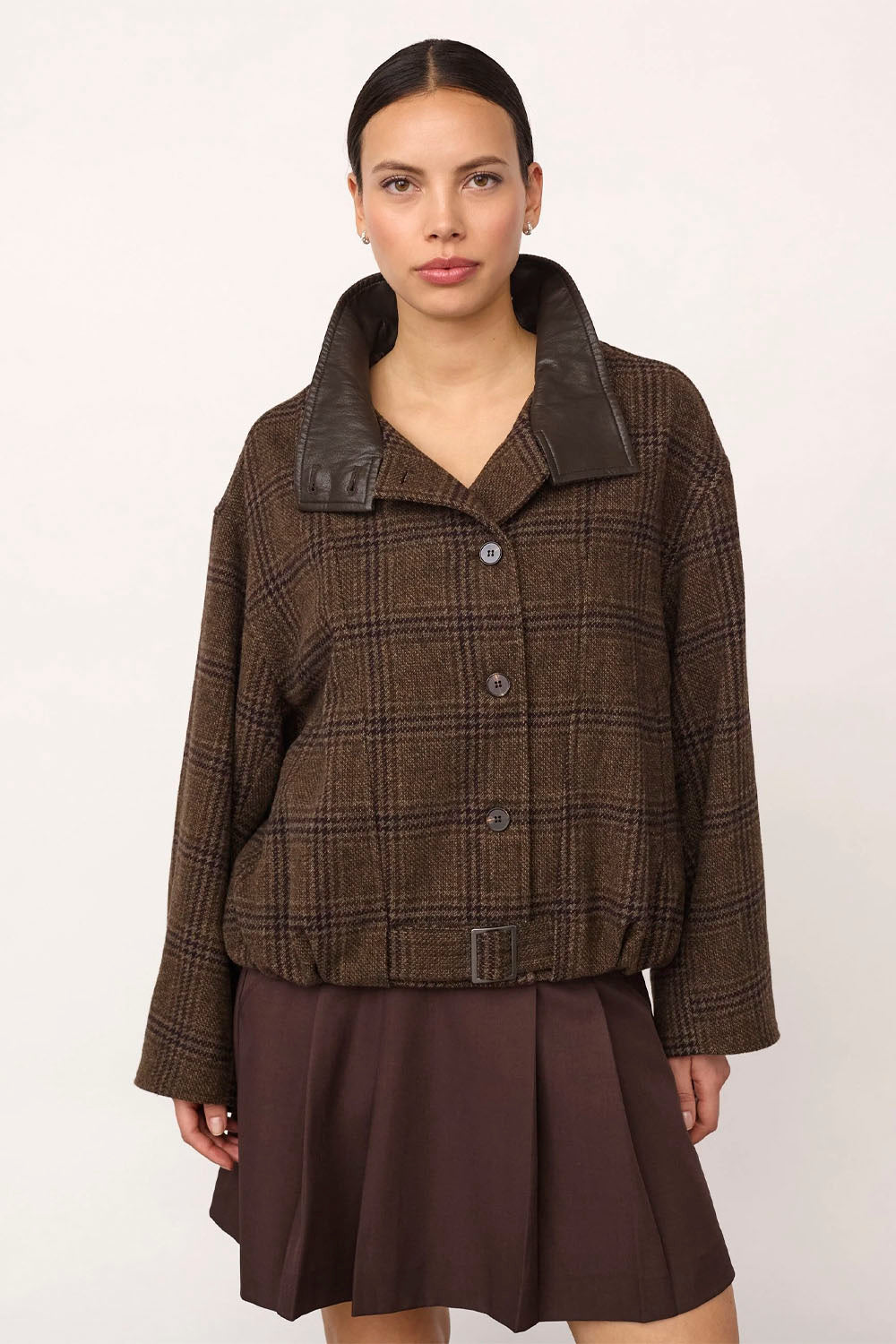 A model wearing a brown and black plaid cropped jacket with dark brown faux-leather lapels and a matching dark brown pleated skirt, photographed against a plain off-white background.