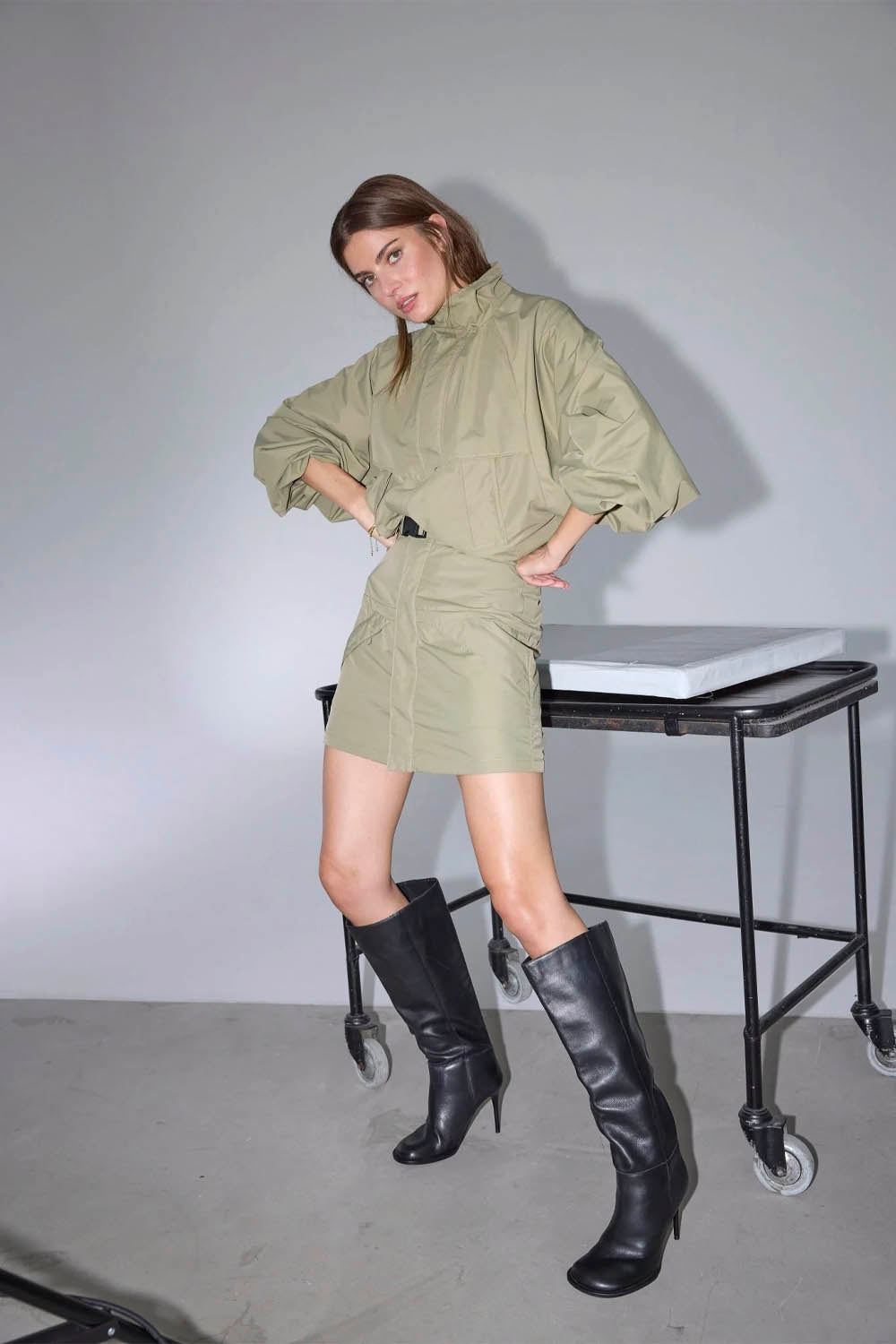 Woman in a green outfit with black knee-high boots standing next to a table.