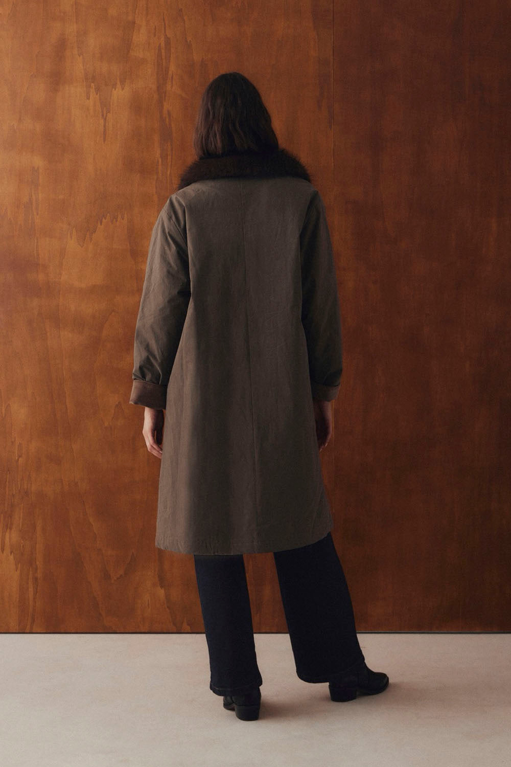 A person viewed from behind wearing a long dark olive-brown coat with a dark fur collar, paired with black wide-leg trousers and black shoes, standing against a warm brown textured wall.