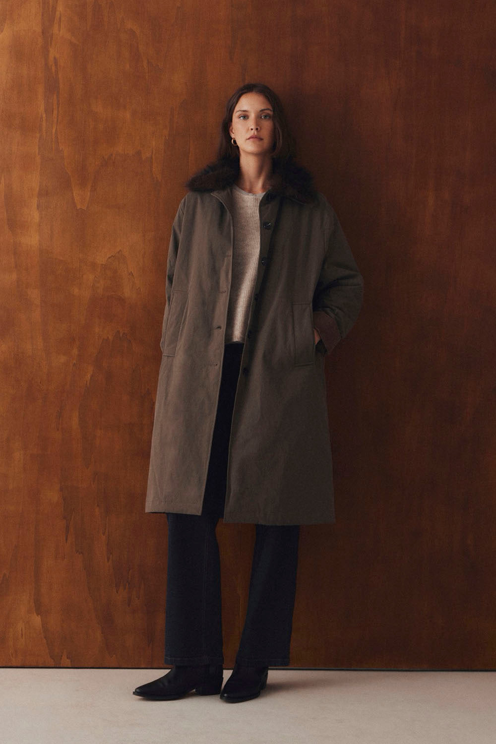 A woman stands against a warm brown wooden wall wearing a long taupe-brown coat with dark fur trim, a light beige top, dark wide-leg trousers, and black shoes.