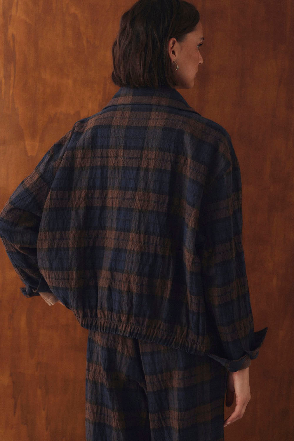 A model shown from the back wearing a dark blue and brown plaid jacket and matching pants against a warm brown textured background.