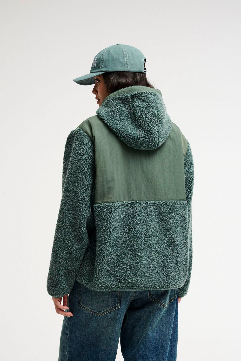 A person shown from the back wearing a green hooded fleece jacket with a solid green back panel, blue jeans, and a matching green cap against a plain light gray studio background.