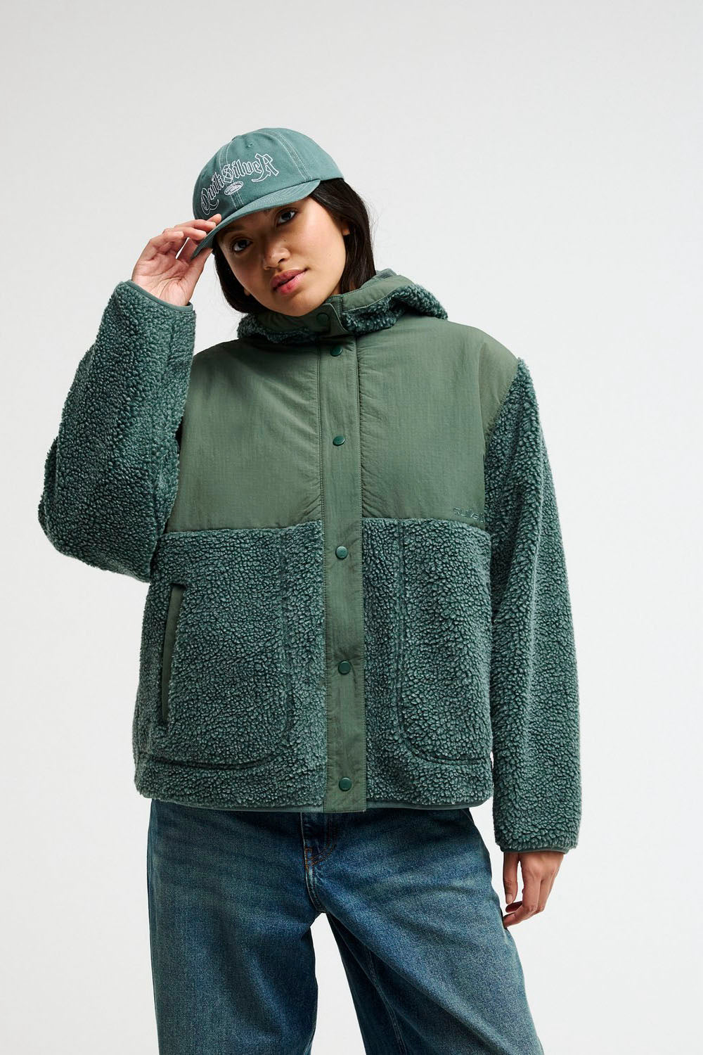 A model wearing a green color-blocked fleece jacket with a hood, paired with blue jeans and a matching green cap, against a plain light gray background.