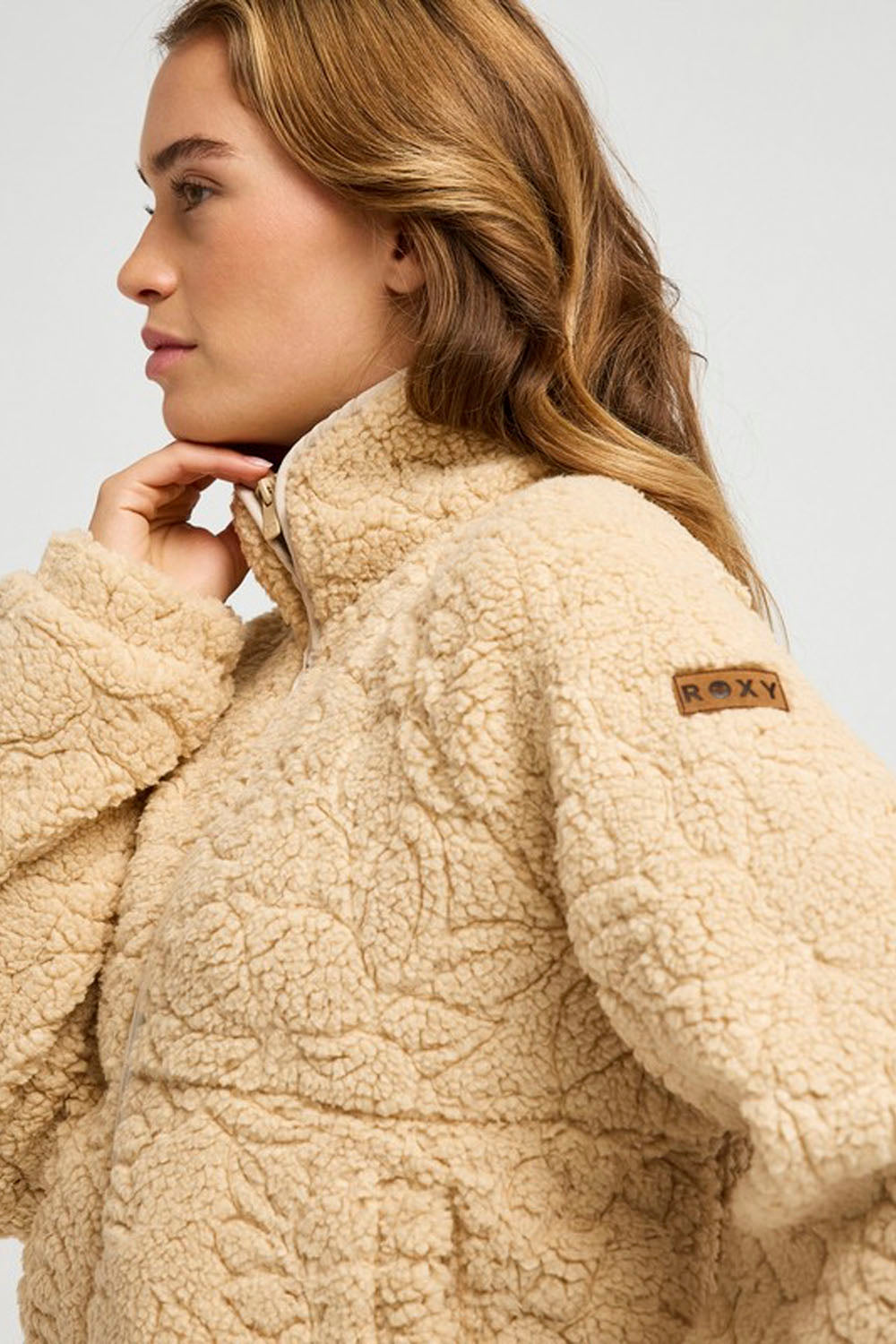 A close-up of a woman wearing a beige sherpa fleece jacket with a textured pattern and a small brown Roxy brand patch on the sleeve, set against a plain light gray background.