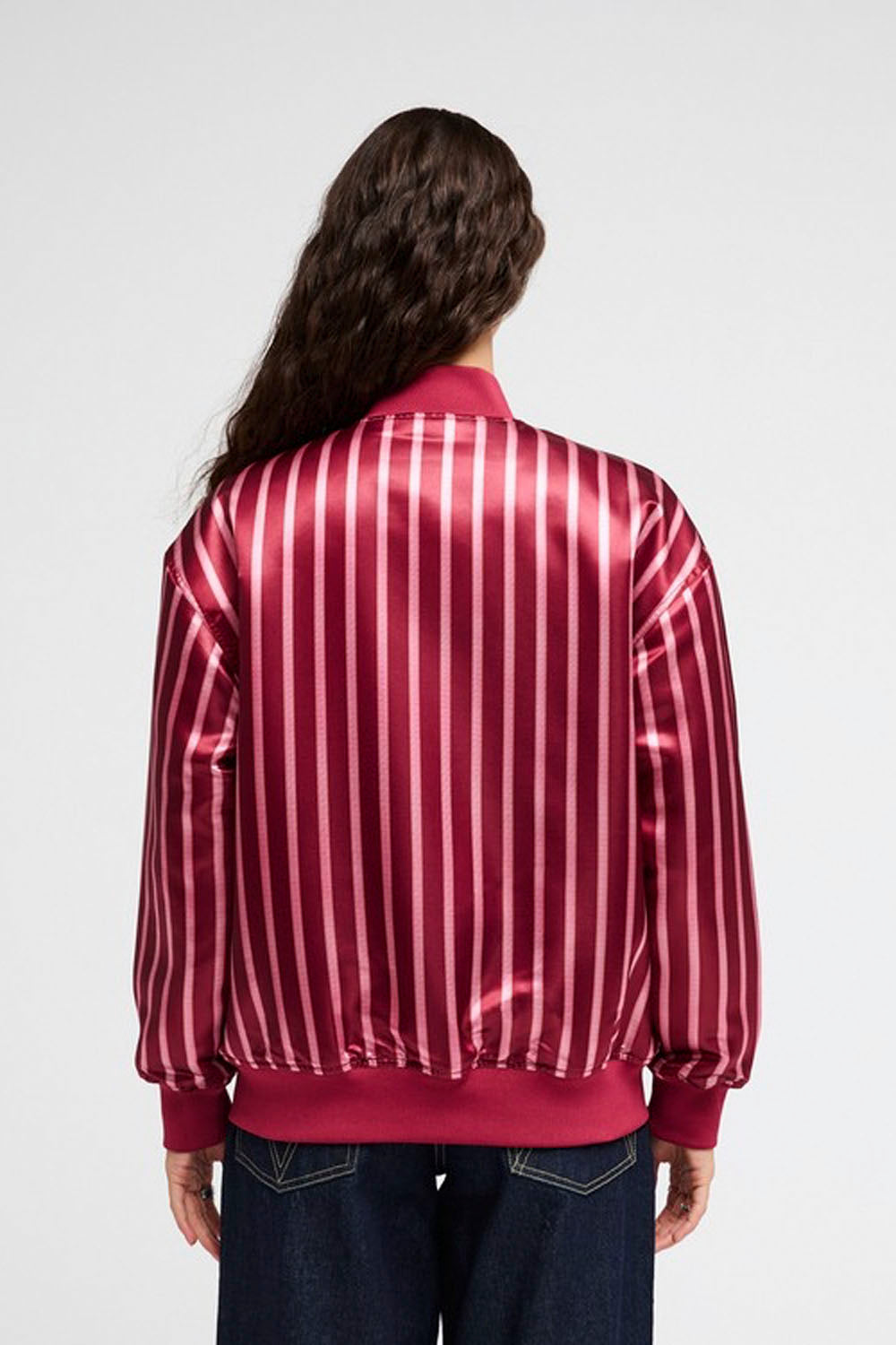A model viewed from the back wearing a shiny satin bomber jacket with vertical pink and white stripes, paired with dark blue jeans, against a light gray studio background.