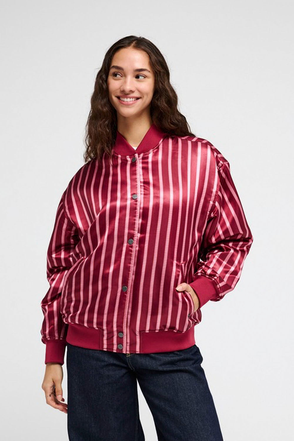 A woman wearing a shiny satin bomber jacket with vertical red and white stripes, paired with dark blue jeans, photographed against a light gray studio background.