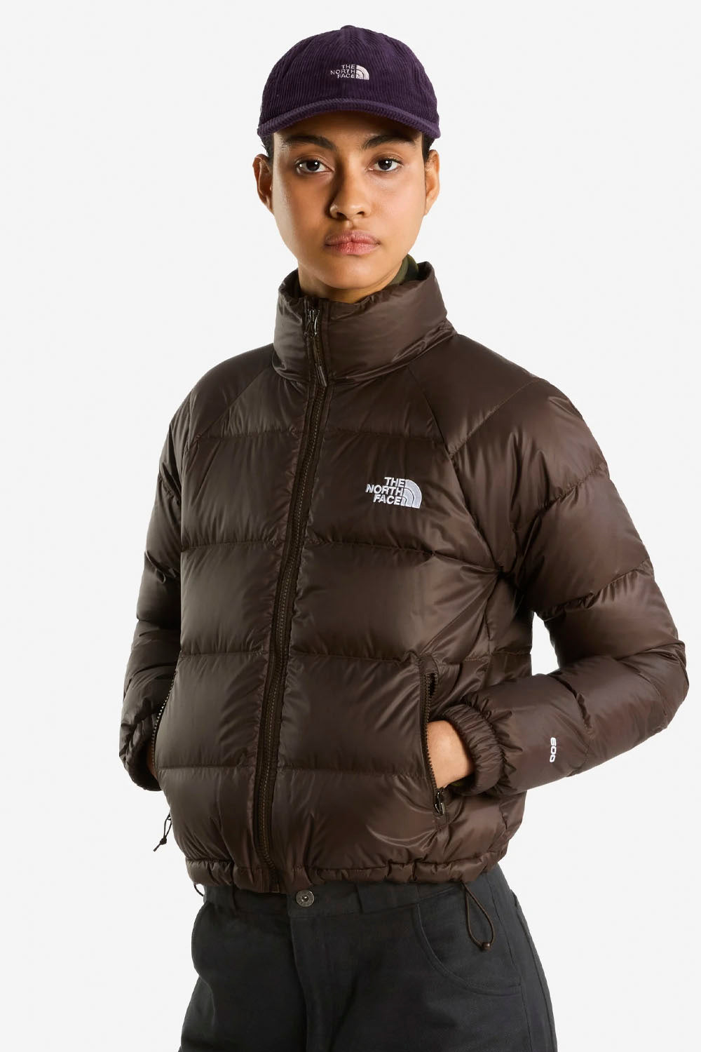 A model wearing a dark brown puffer jacket by The North Face, paired with a matching dark cap and dark trousers, against a plain light gray background.