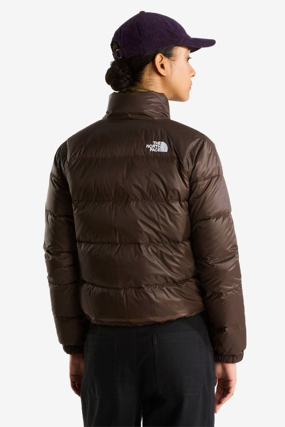 A person shown from the back wearing a dark brown puffer jacket with a white The North Face logo on the upper back, paired with black pants and a dark purple cap, against a light gray studio background.