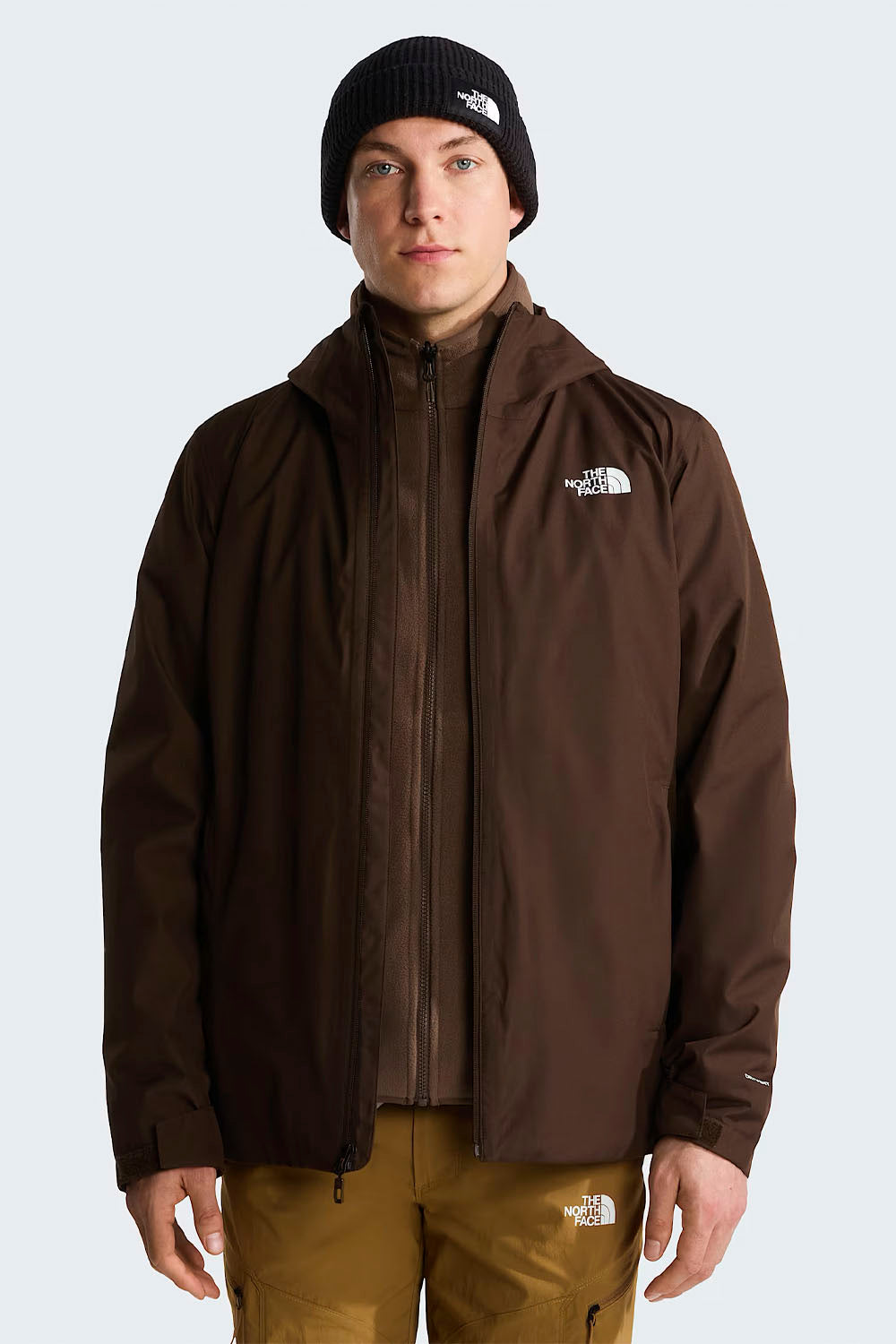 A man wearing a dark brown hooded jacket by The North Face, shown from the back. He is also wearing a black beanie and tan pants against a light gray studio background.