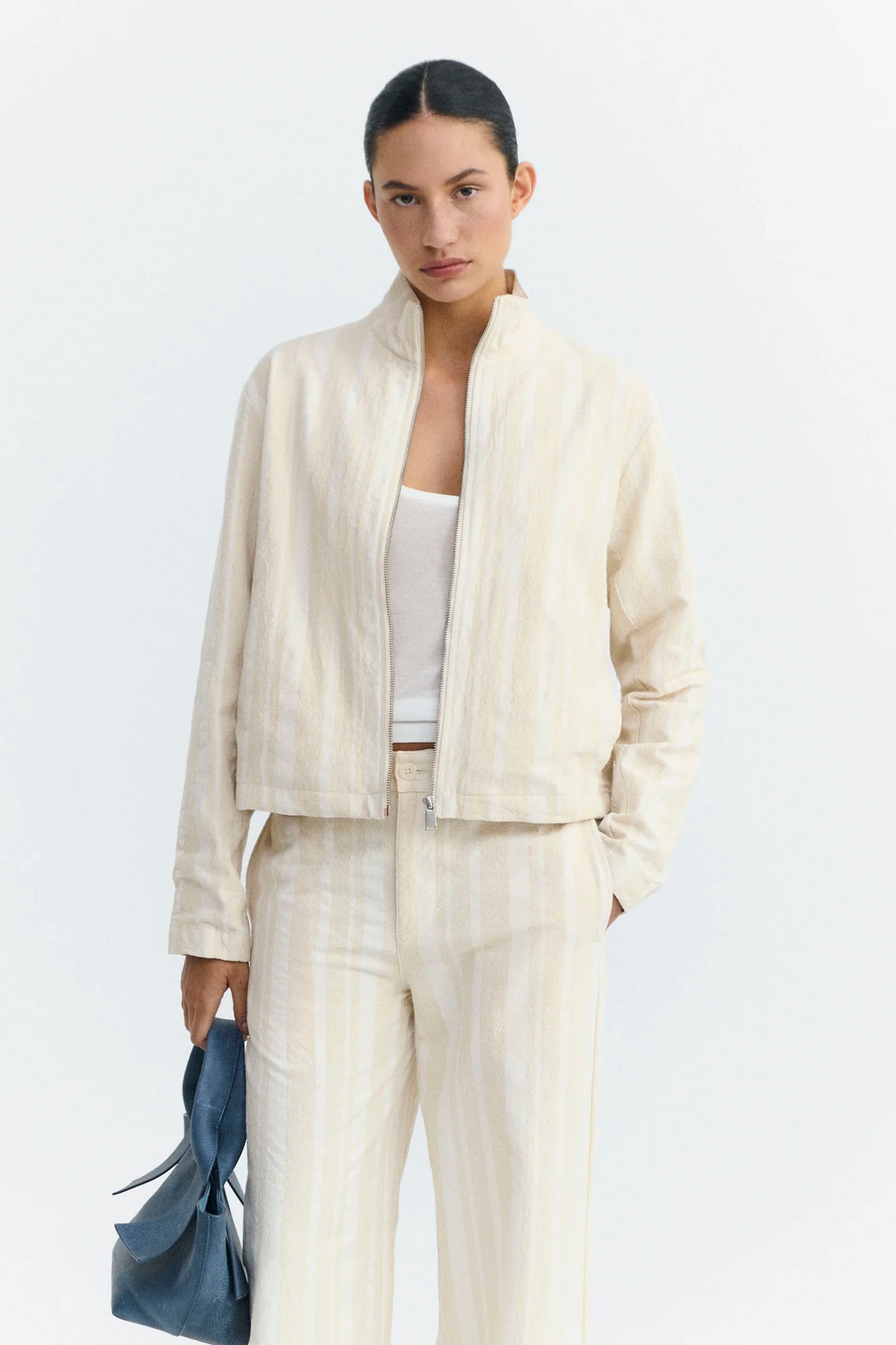 Woman wearing a cream Thinking Mu jacket and holding a blue bag on a light gray background