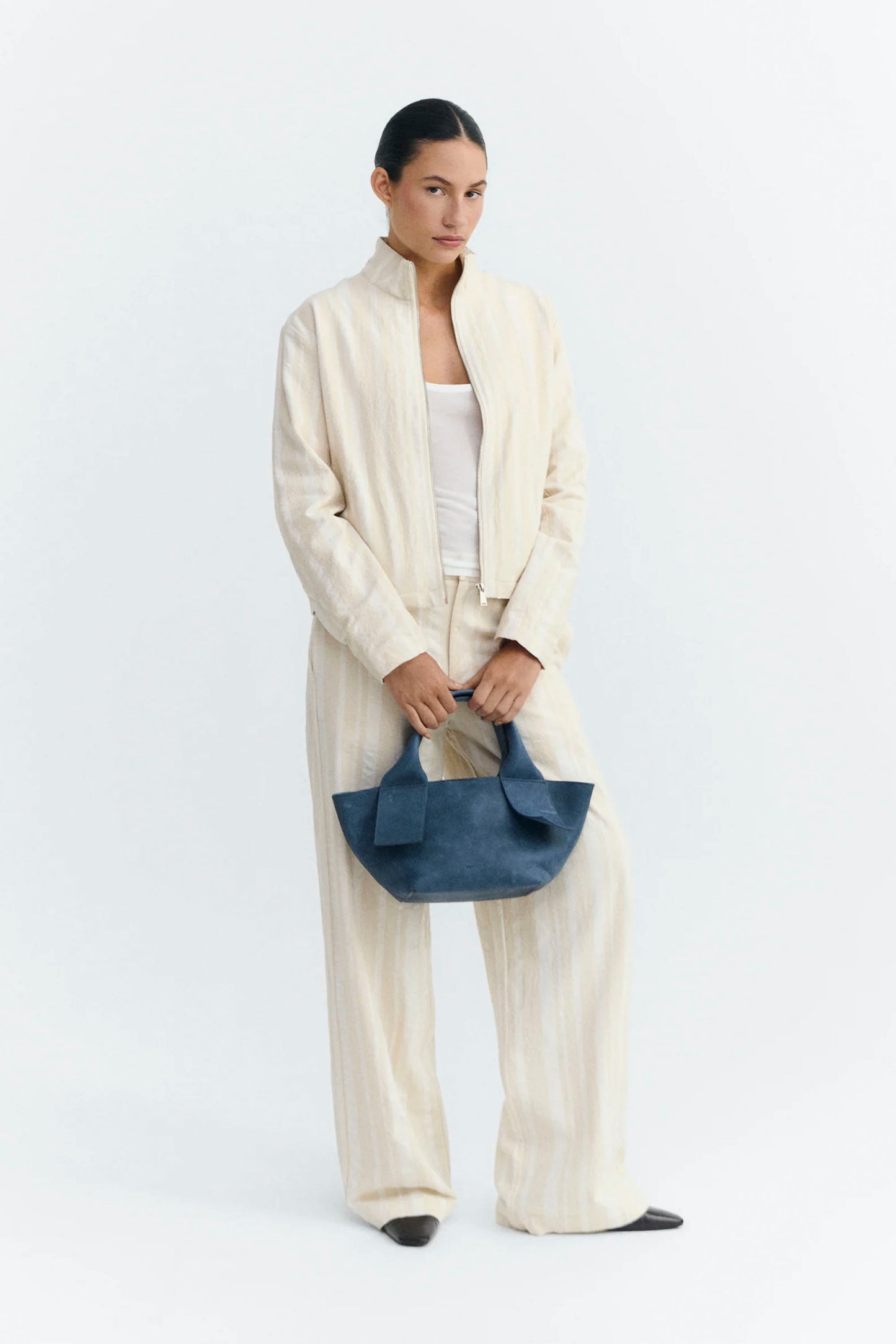 Woman holding a blue handbag wearing a cream Thinking Mu jacket on a white background
