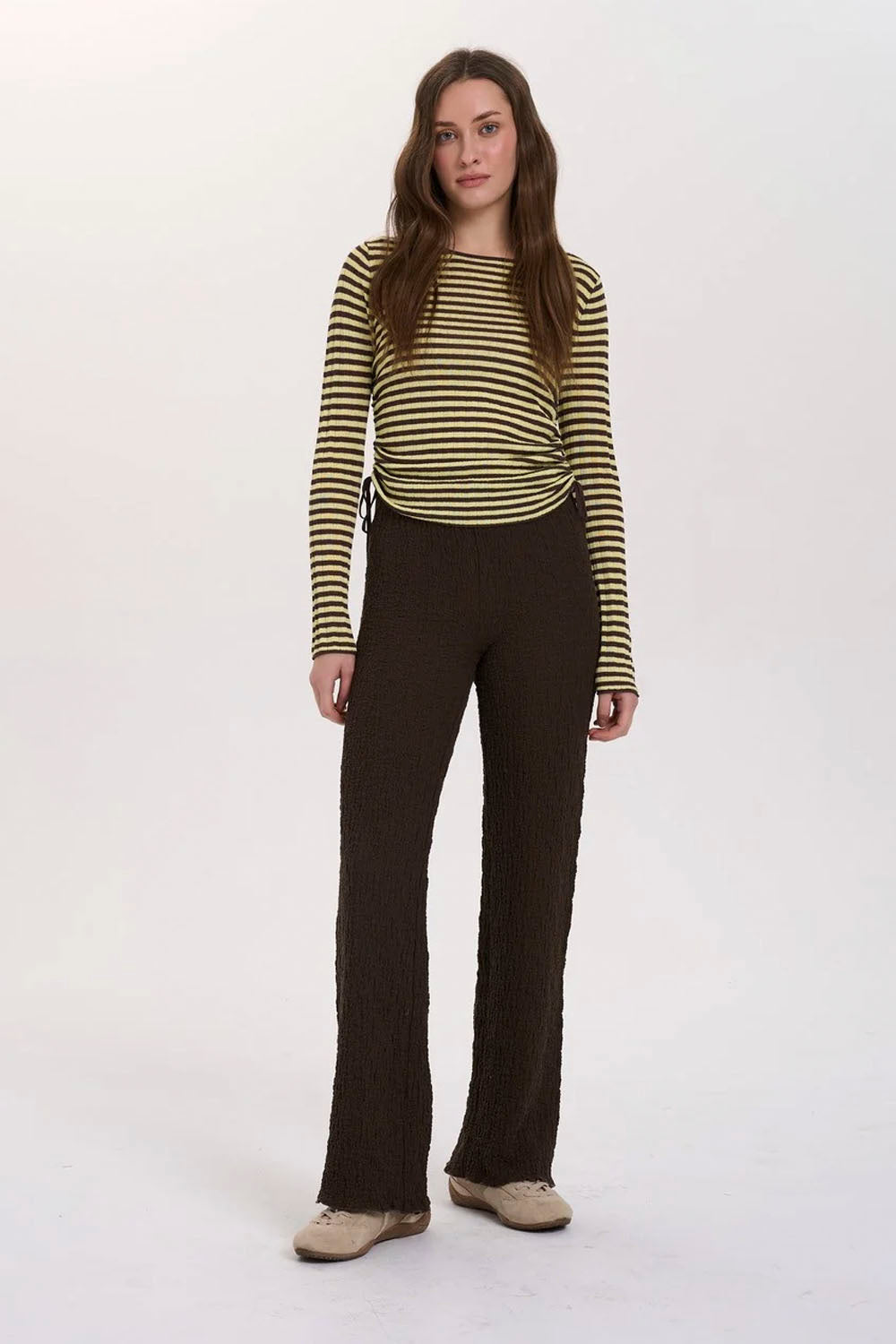 A woman modeling a long-sleeve knit top with horizontal olive green and beige stripes, paired with dark brown flared pants and beige shoes, against a plain white studio background.