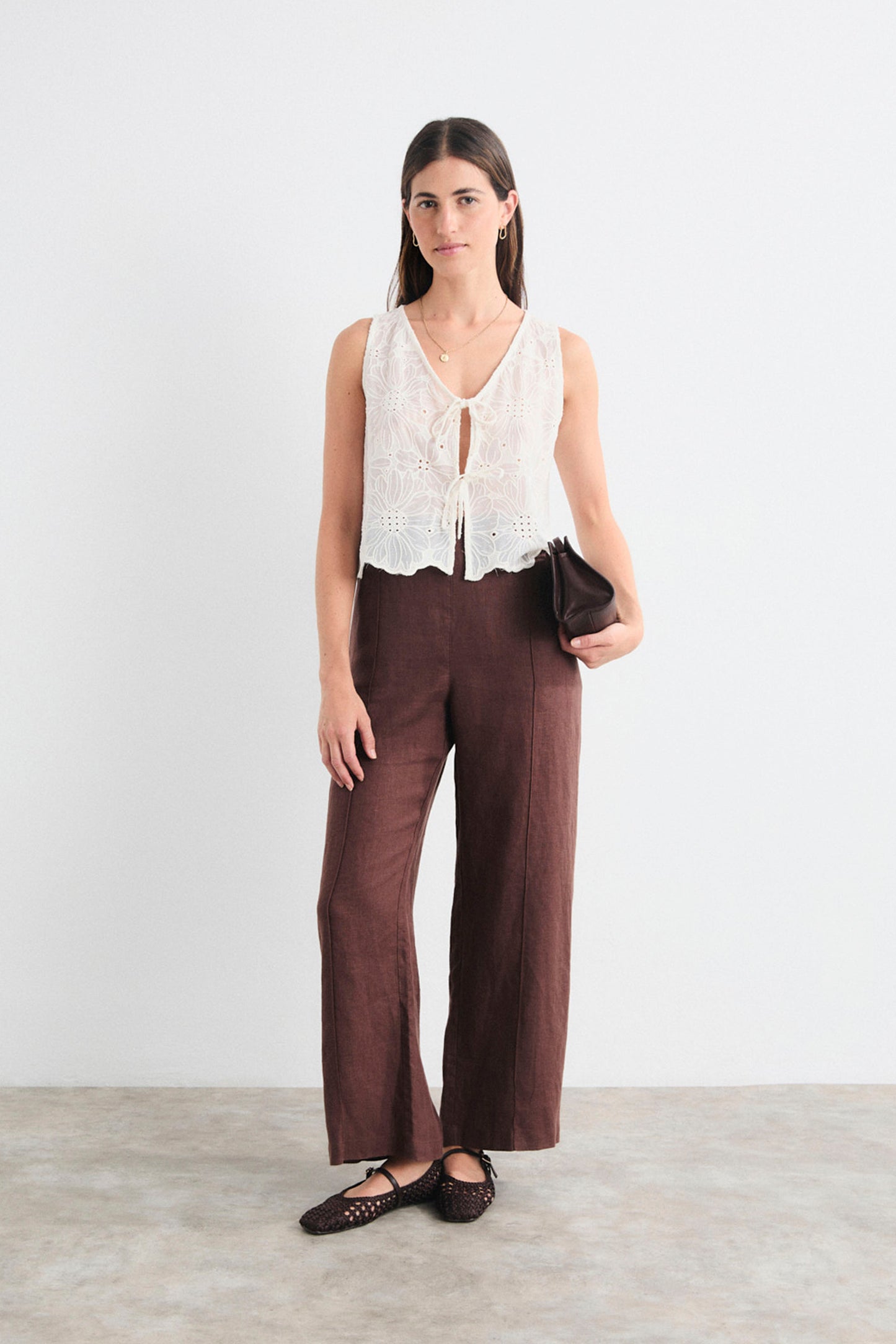 Woman wearing a white sleeveless top with lace details and brown pants against a white background