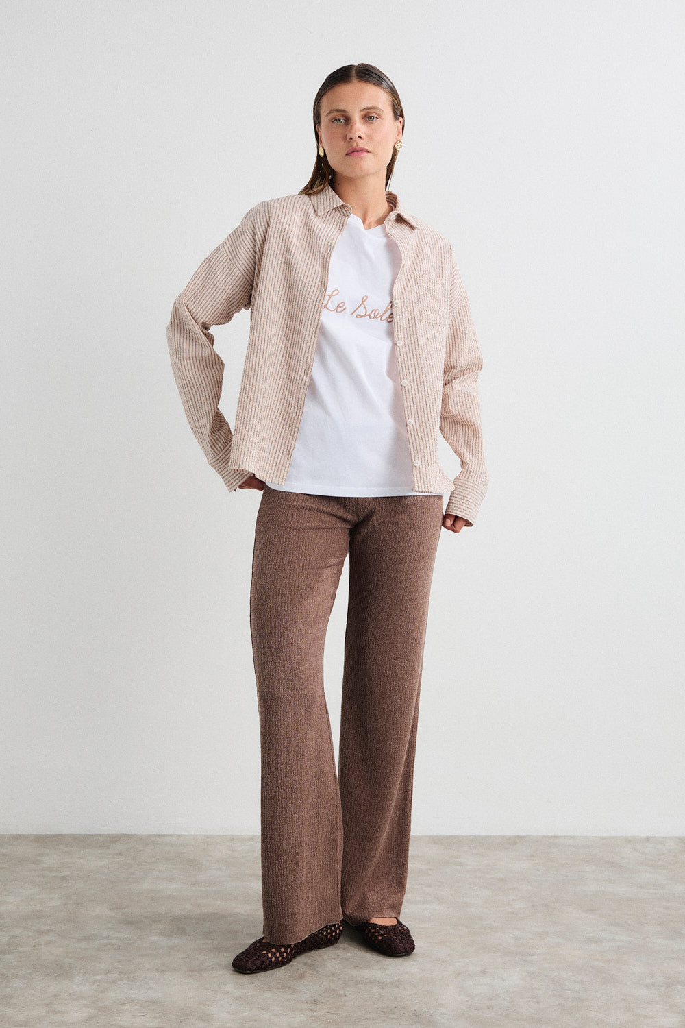 Woman wearing a beige jacket, white shirt, and brown pants on a plain background