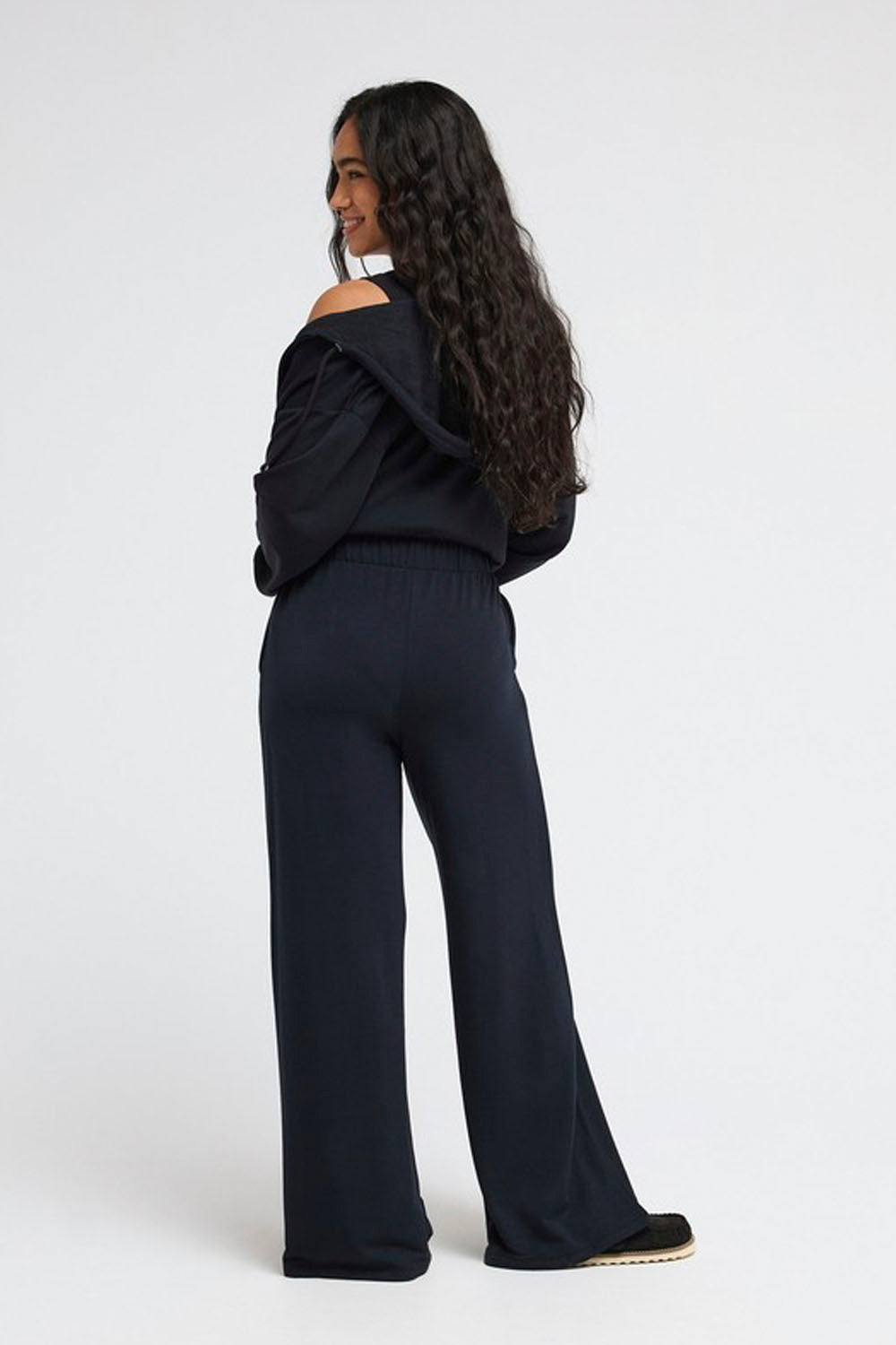 A woman wearing a dark navy off-the-shoulder long-sleeve top and matching wide-leg trousers, shown from the back against a plain light gray studio background.