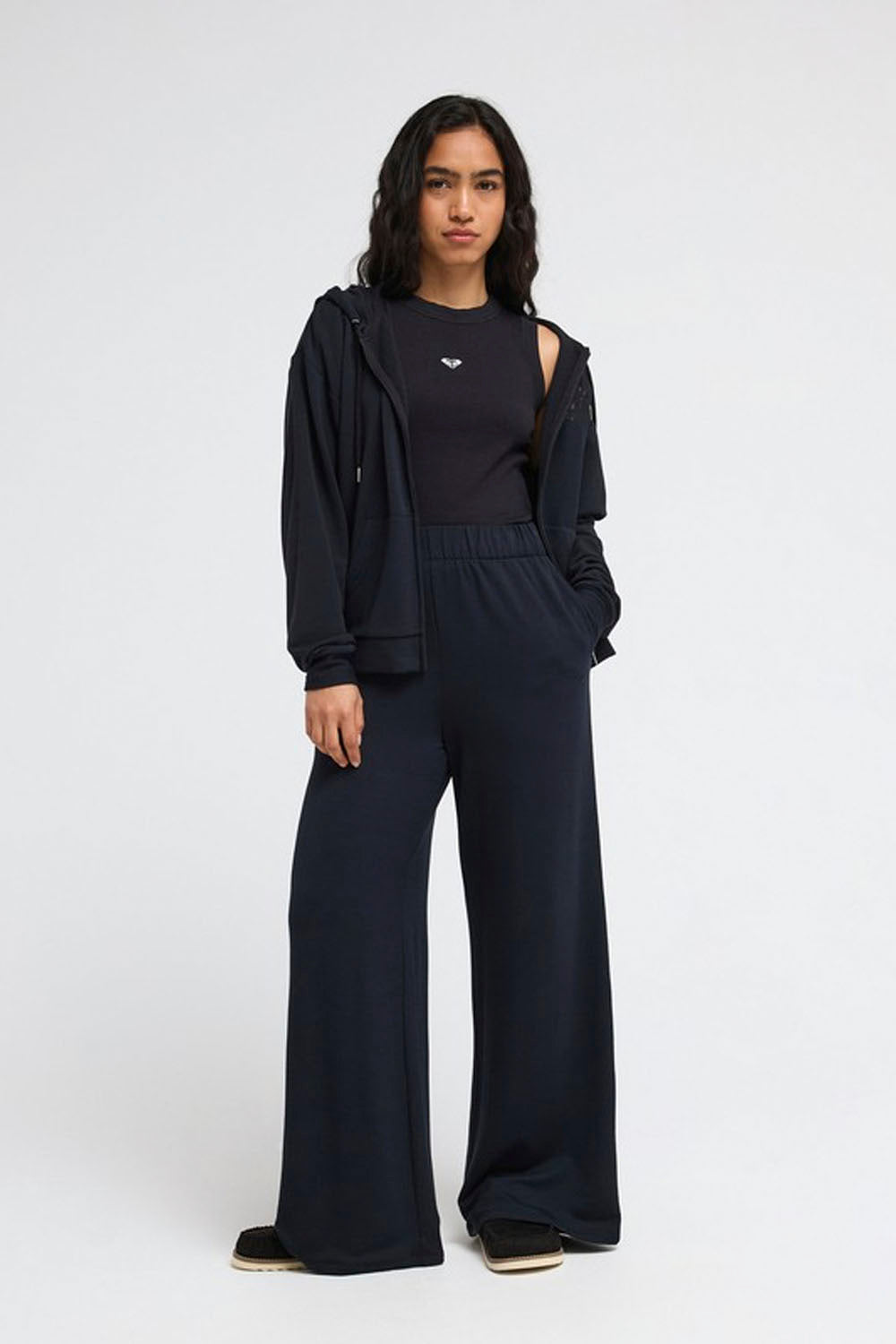 A model stands against a light gray studio background wearing a dark navy blue coordinated outfit consisting of a zip-up hoodie, a short-sleeve top with a small white logo, and wide-leg pants.