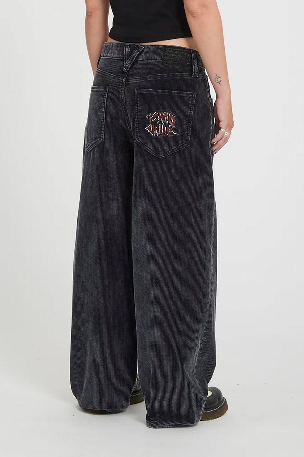 A person shown from the back wearing very wide-leg black denim jeans with a small embroidered graphic on the back pocket, paired with a black top and black shoes, against a plain light gray studio background.