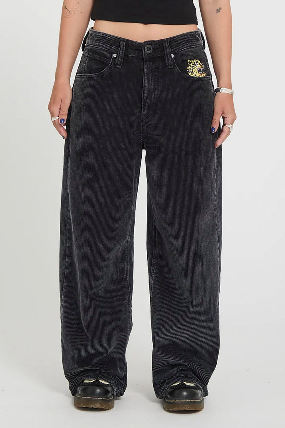 A person wearing oversized faded black denim jeans with a small gold embroidered patch on the pocket, shown from the waist down against a light gray studio background.