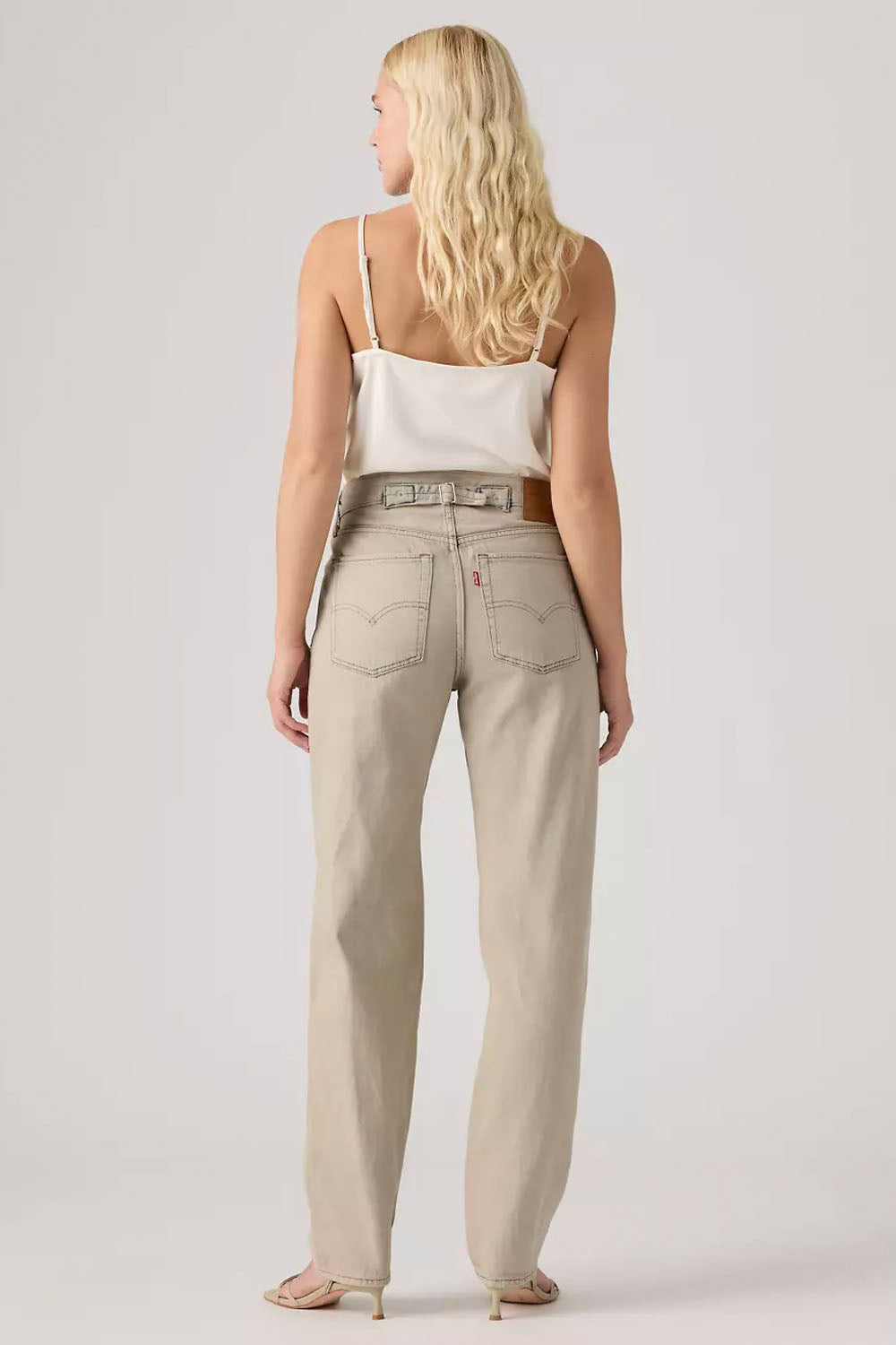 A woman shown from the back wearing light beige straight-leg jeans with a silver-toned belt, a white camisole top, and beige high-heeled sandals, photographed against a light gray studio background.