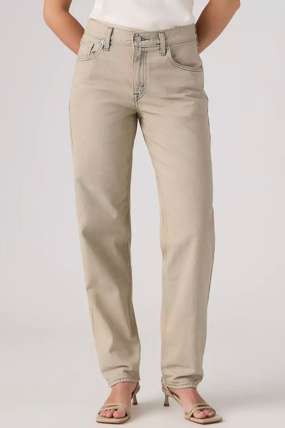 A person wearing light beige straight-leg denim jeans with a white top and beige heeled sandals, shown from the waist down against a light gray studio background.