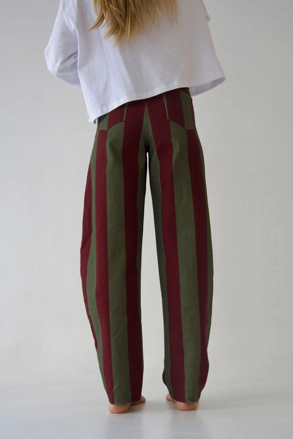 A person viewed from behind wearing loose, wide-leg striped pants in dark red and olive green, paired with a white cropped top, against a plain light gray studio background.