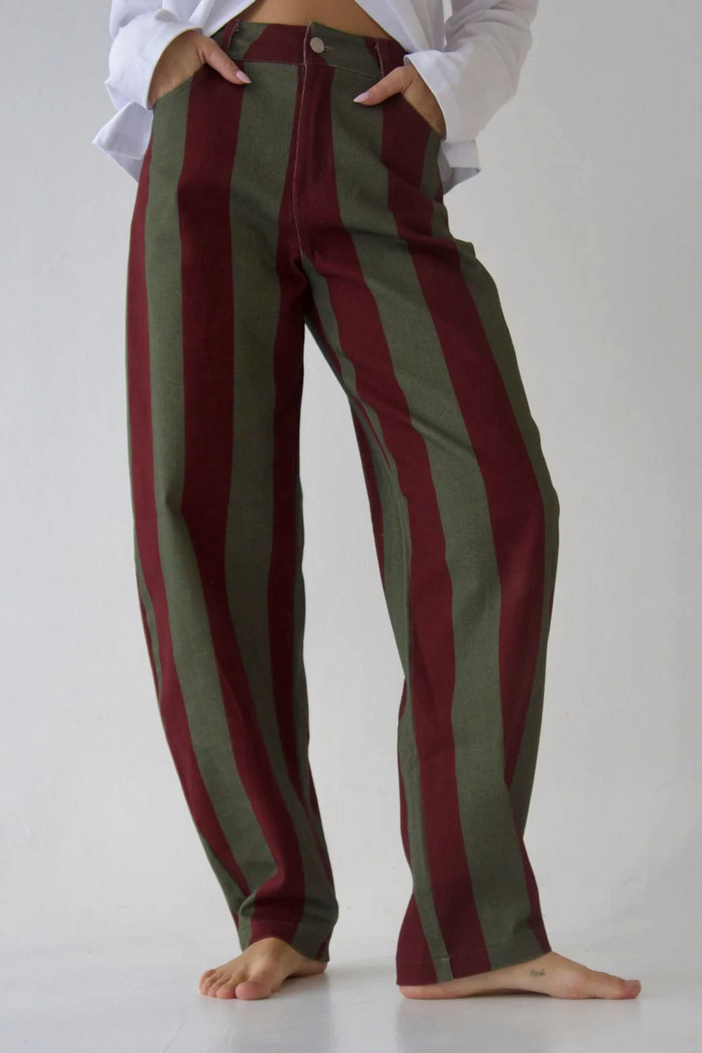 A person wearing loose, wide-leg pants with vertical dark red and olive green stripes, paired with a white shirt, standing barefoot against a light gray studio background.