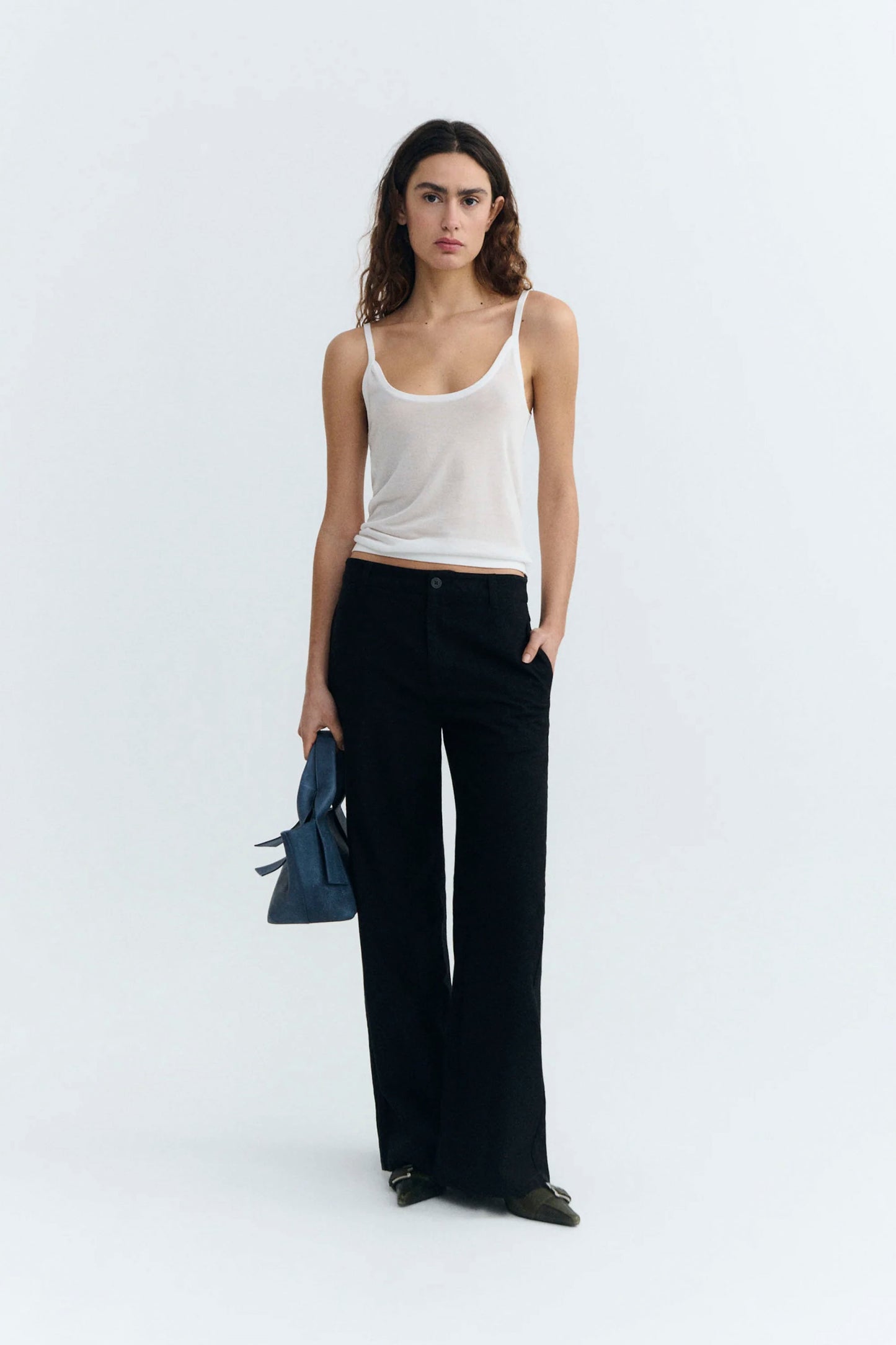 Woman wearing a white tank top and black Thinking Mu pants holding a blue clutch against a white background