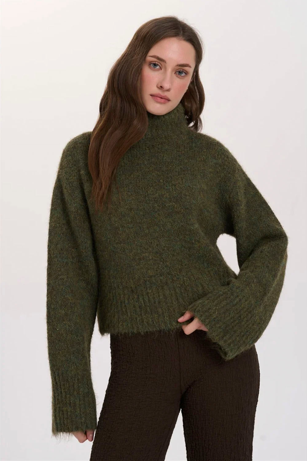A woman wearing a fuzzy olive green mock-neck knit sweater and dark brown pants, photographed against a plain light background.