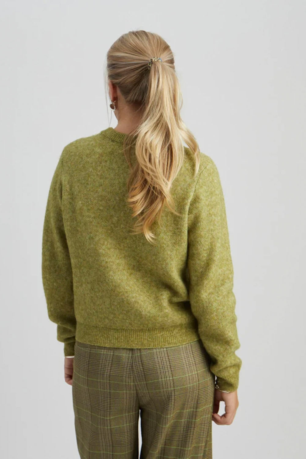 A woman viewed from behind wearing a textured olive green knit sweater and olive plaid trousers, set against a plain light gray studio background.