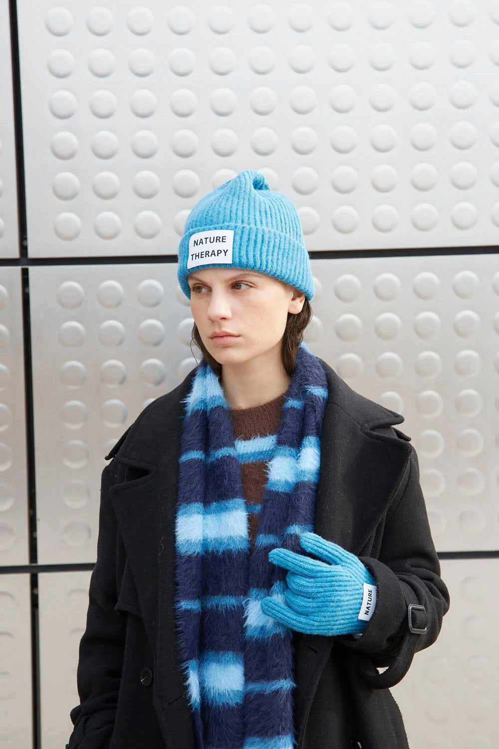 A model wearing a light blue knit beanie with a 'NATURE THERAPY' label, a blue and navy striped fuzzy scarf, a light blue knit glove, and a black coat, standing against a textured gray wall.