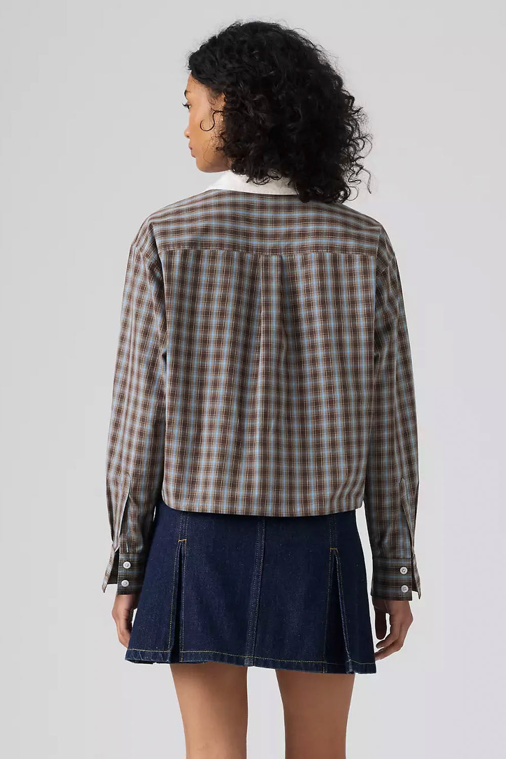 A model shown from the back wearing a brown and beige plaid button-up shirt with a white collar, paired with a dark blue denim pleated skirt, against a light gray studio background.