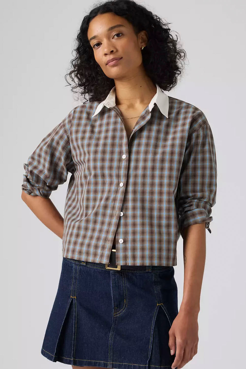 A woman wearing a brown and beige plaid button-down shirt with rolled-up sleeves and a dark blue denim mini skirt, photographed against a light gray studio background.