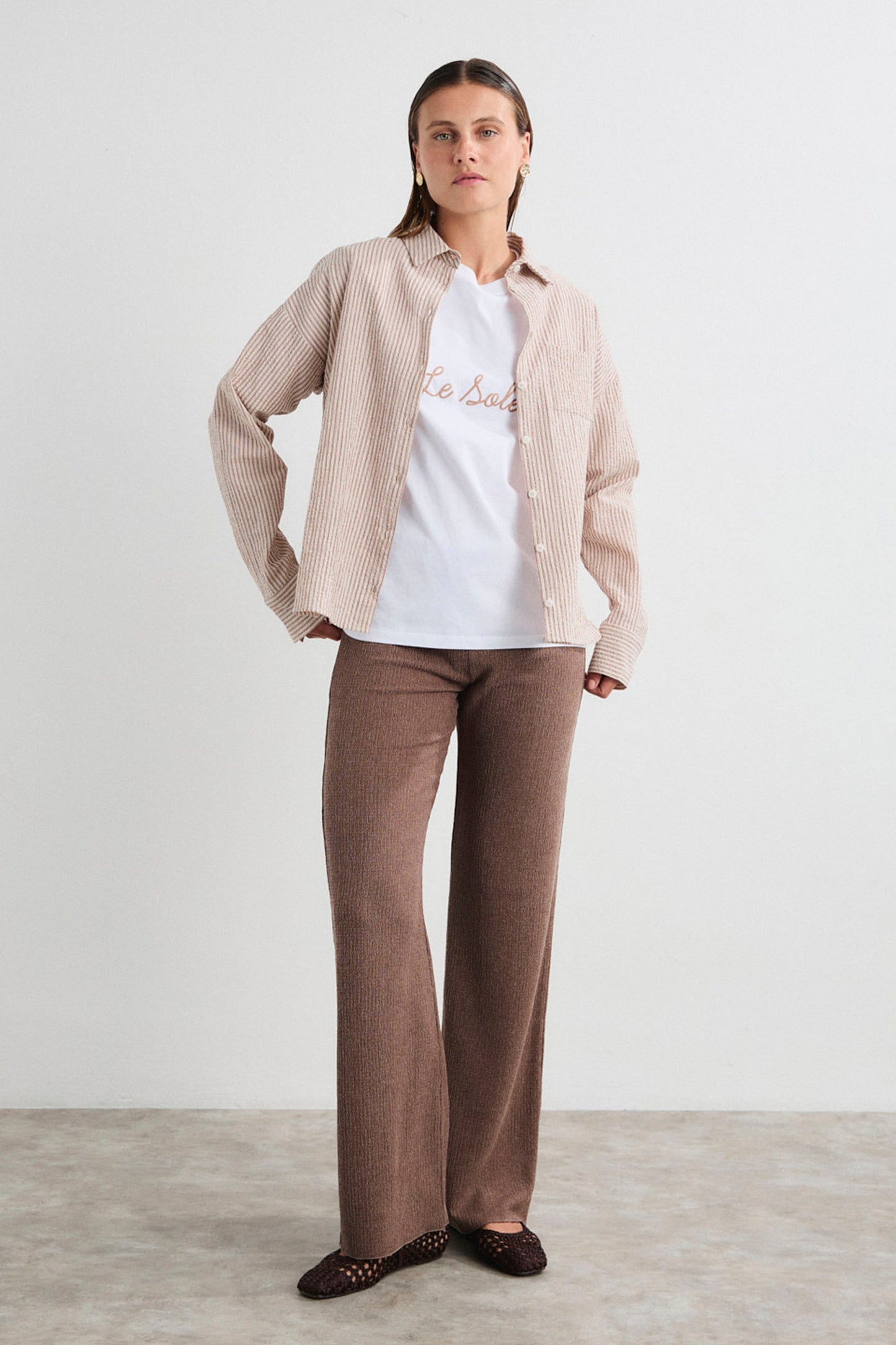 Woman wearing a beige shirt, white t-shirt with text, and brown pants on a plain background