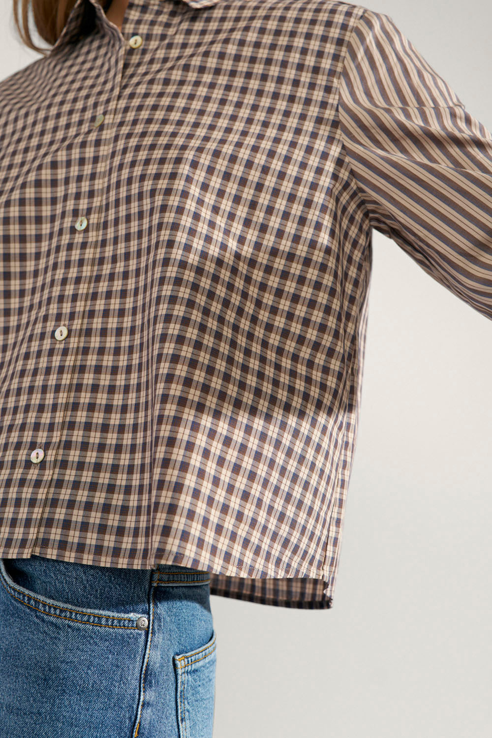 A cropped long-sleeve button-up shirt with a brown, beige, and white checkered pattern, styled with blue jeans against a light gray background.