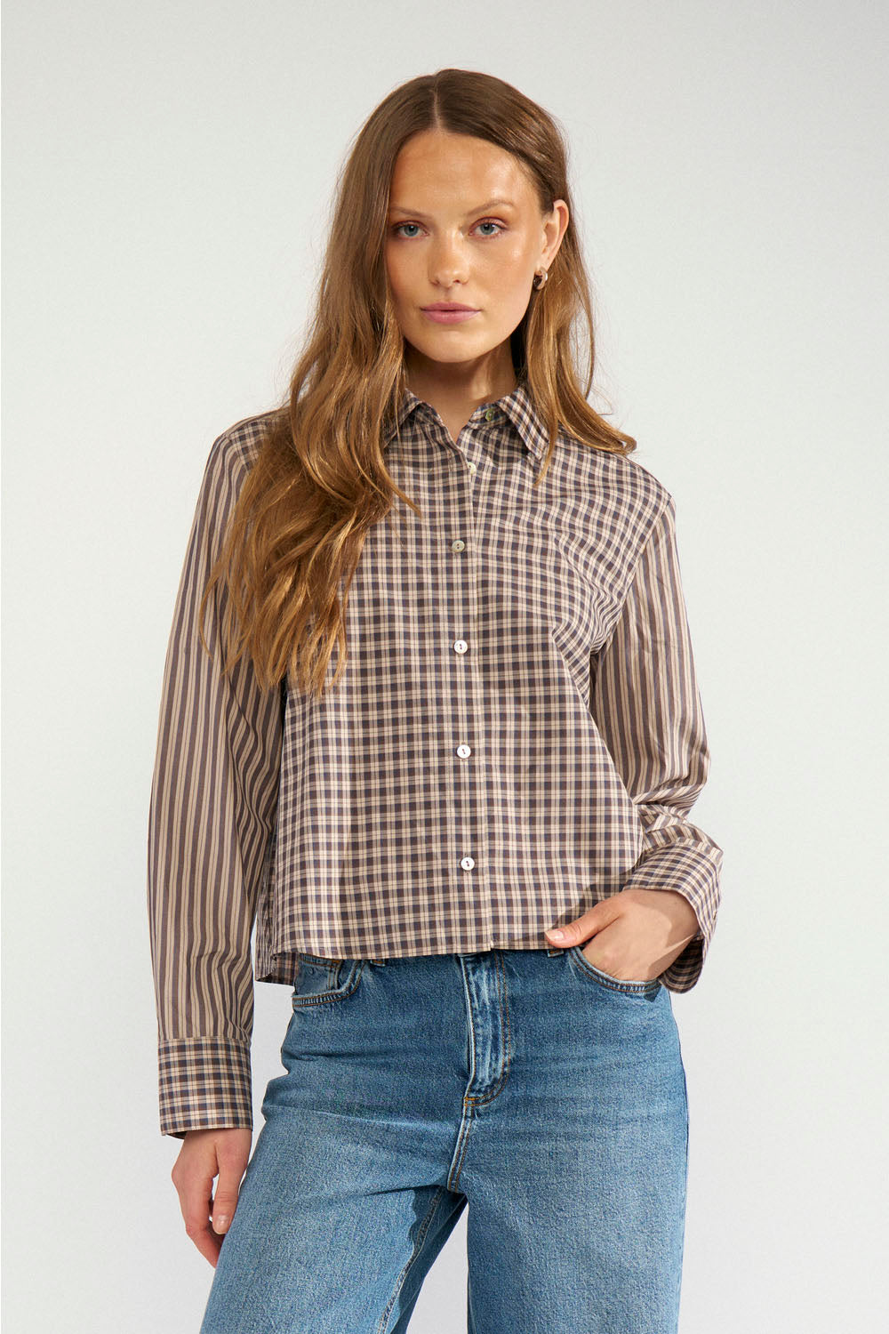 A woman modeling a long-sleeve button-up shirt with a brown and beige check pattern, paired with blue jeans, against a light gray studio background.