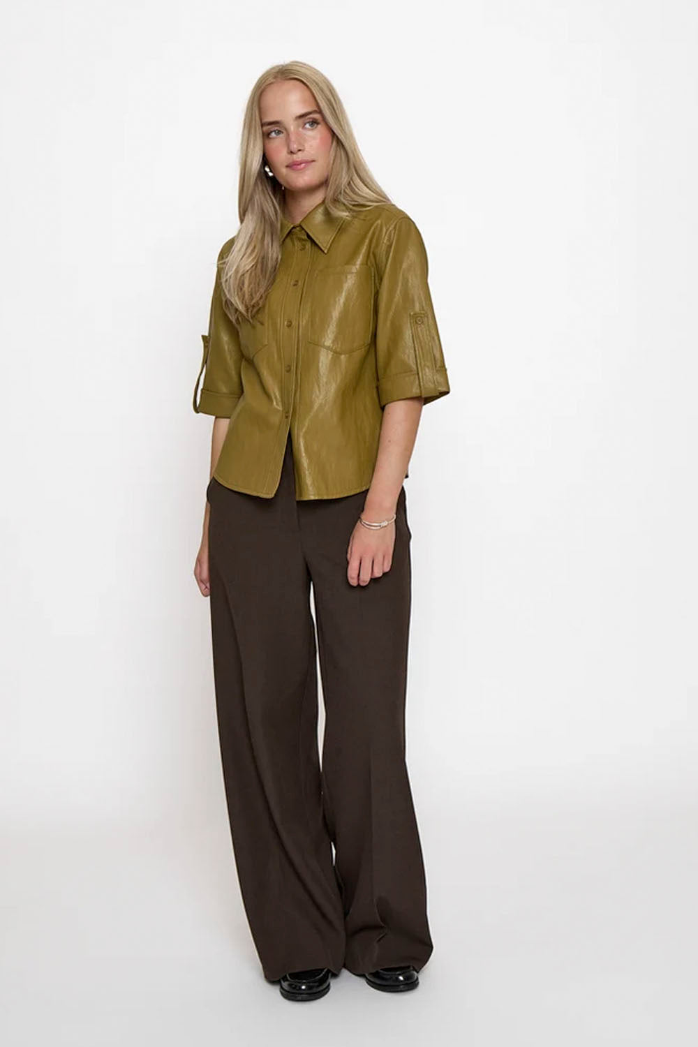 A woman modeling a short-sleeved olive green faux-leather button-up shirt paired with dark brown wide-leg trousers, standing against a plain white studio background.