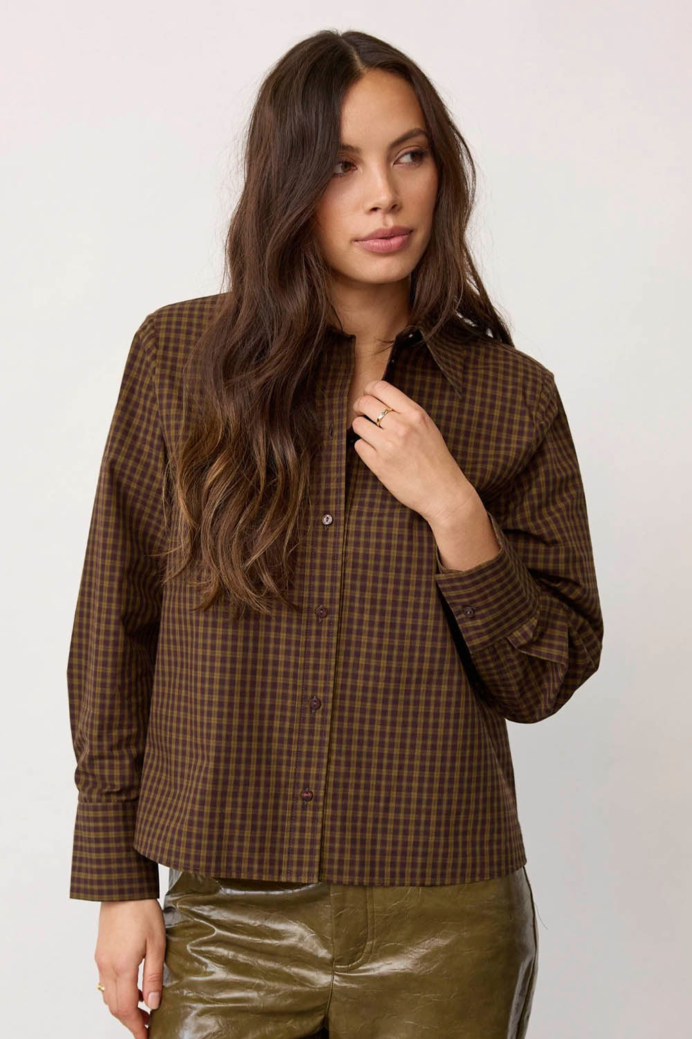A woman wearing a brown and black checkered button-down shirt with olive green pants, posing against a light gray studio background.