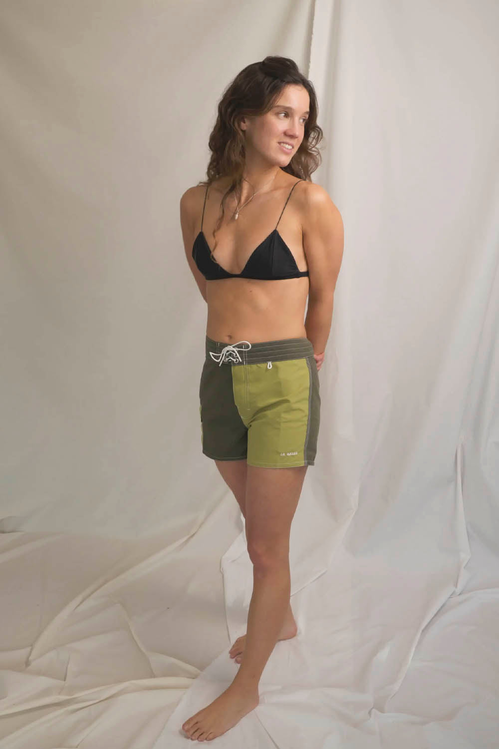 A woman modeling a pair of olive green and black swim shorts with a black triangle bikini top, standing against a draped white fabric backdrop.