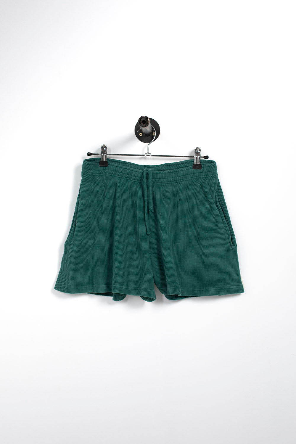 A pair of dark green drawstring shorts hanging from a black metal hanger against a plain white wall.