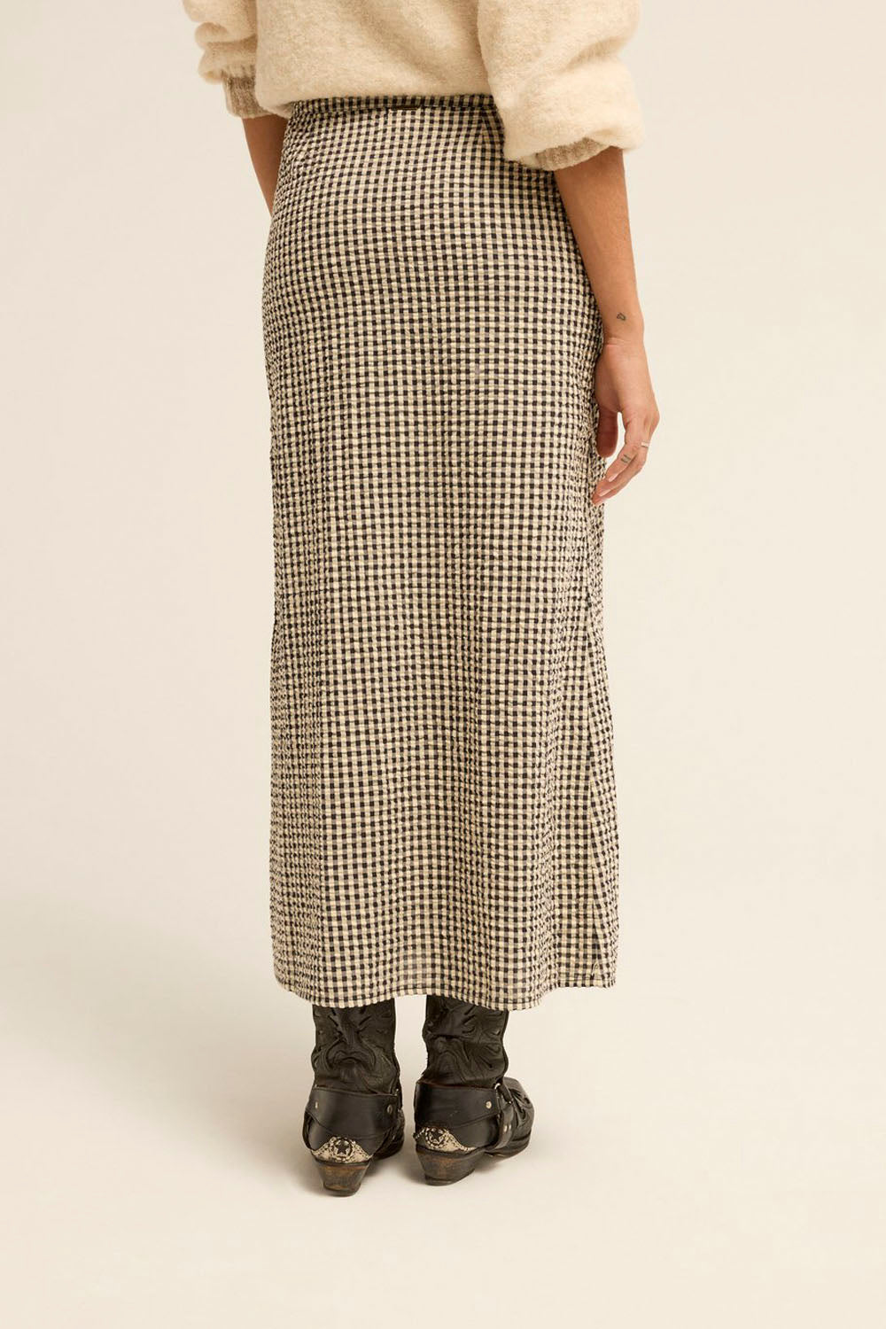 A person shown from the back wearing a long beige and black houndstooth skirt, a cream sweater, and dark boots against a plain light beige studio background.