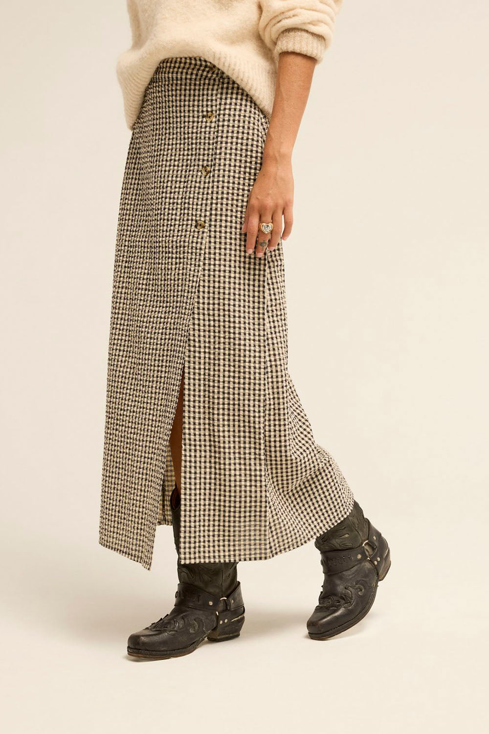 A model wearing a long, wide-leg skirt in a beige and black houndstooth pattern, styled with a cream sweater and black lace-up boots against a warm beige studio background.