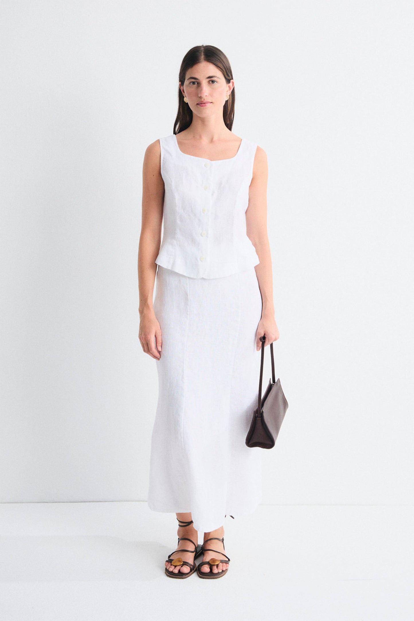 Woman wearing a white sleeveless top and white skirt holding a black handbag on a white background