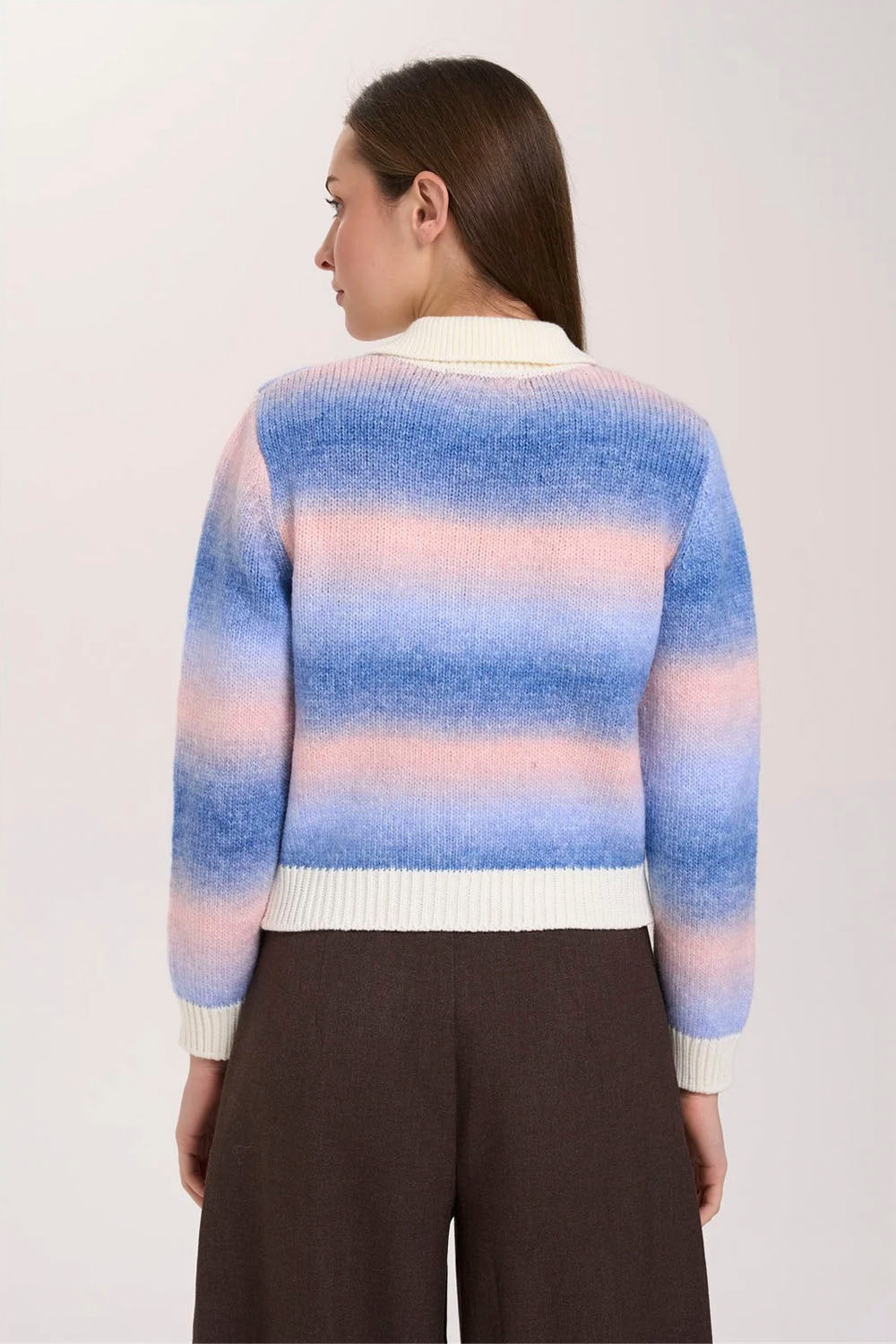 A woman shown from the back wearing a striped knit sweater with horizontal bands of blue, pink, and white, paired with dark brown wide-leg trousers, against a plain light background.
