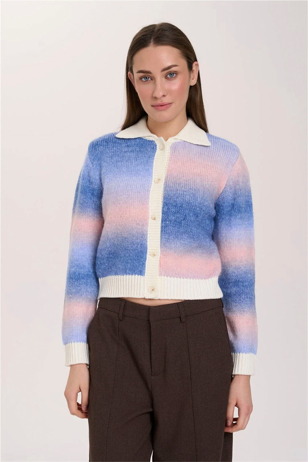 A woman modeling a cropped knit cardigan with a blue, pink, and white gradient pattern, featuring a cream-colored collar and button placket, paired with dark brown trousers against a light gray studio background.