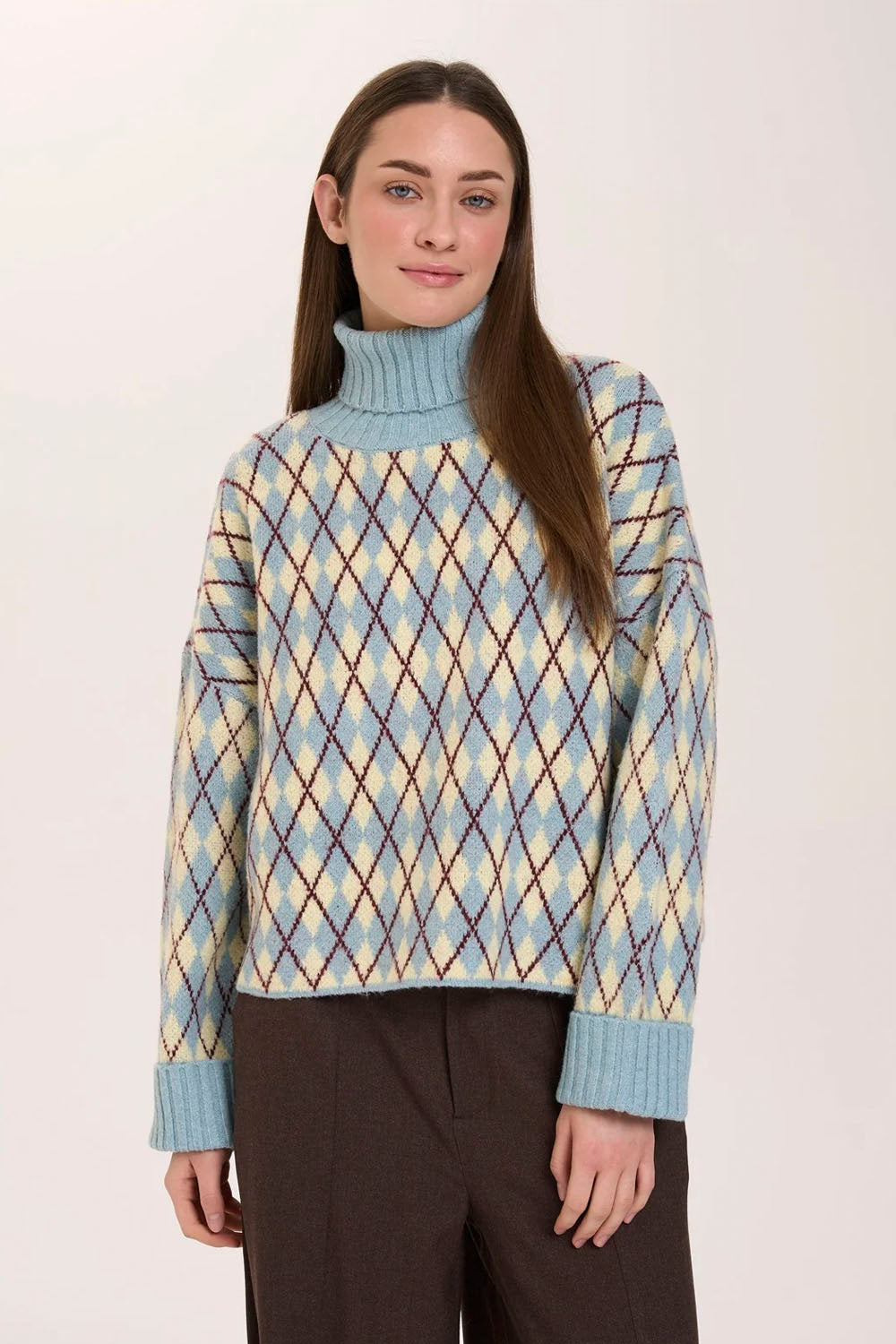 A woman wearing a light blue turtleneck sweater with a yellow and brown argyle pattern, paired with dark brown trousers, against a plain off-white background.