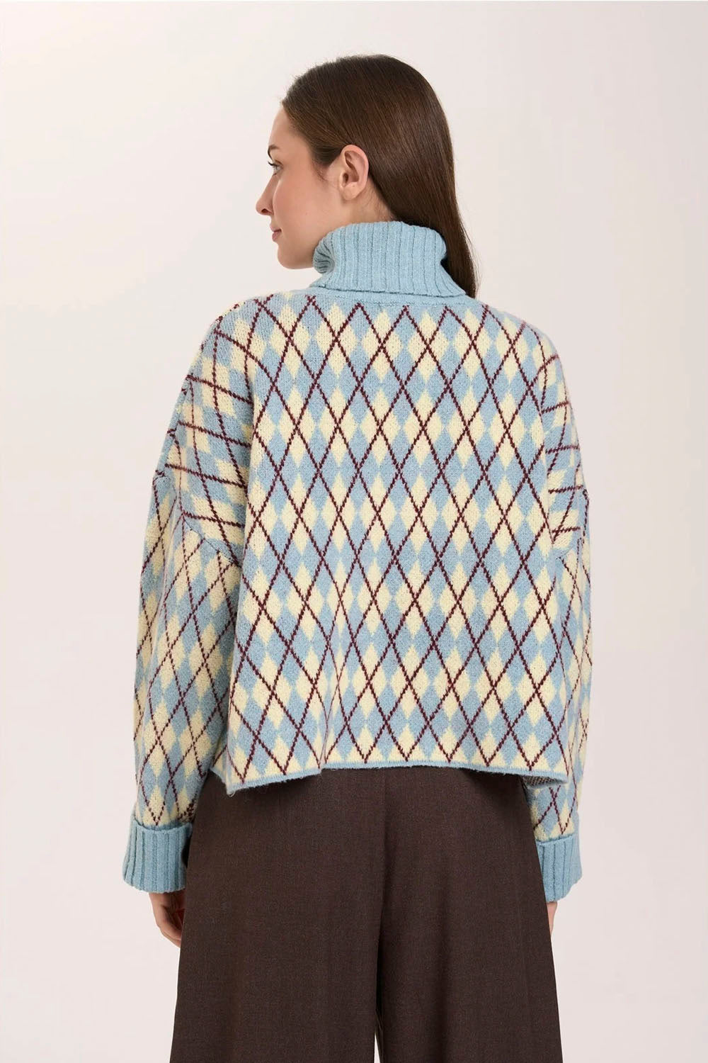 A woman shown from the back wearing a cropped, light blue turtleneck sweater with a beige and brown argyle pattern, paired with dark brown wide-leg trousers, against a plain off-white background.