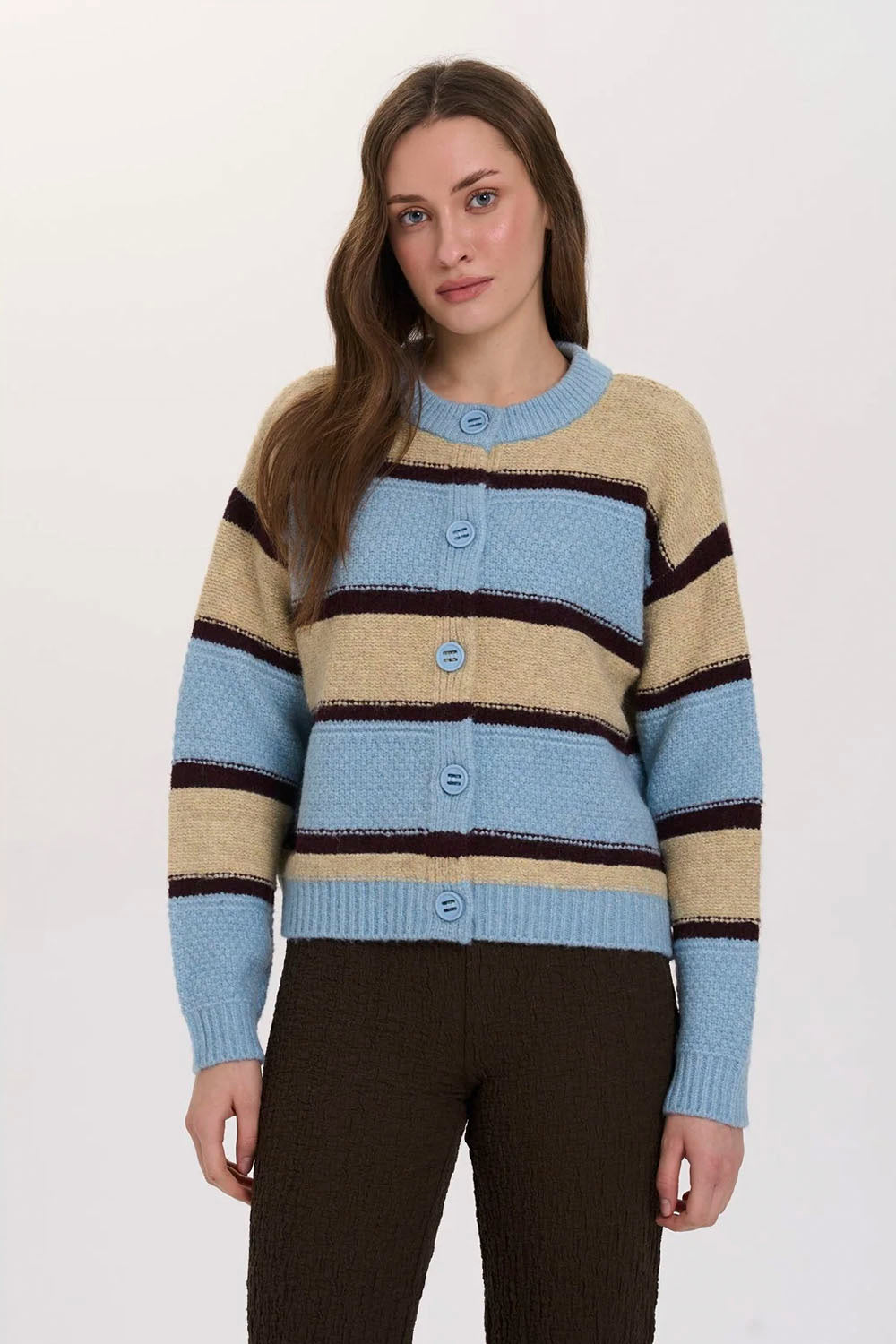 A woman wearing a striped knit cardigan sweater with horizontal bands of light blue, beige, and dark brown, paired with dark brown pants, against a plain light gray studio background.