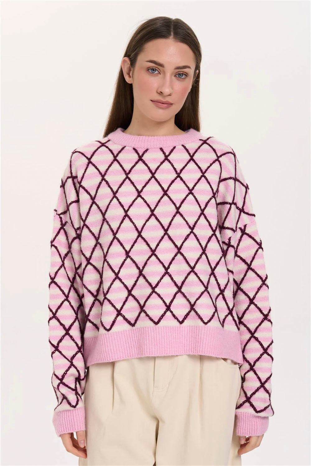 A woman wearing a pink knit sweater with a dark purple diamond lattice pattern, paired with cream-colored pants, against a plain white studio background.