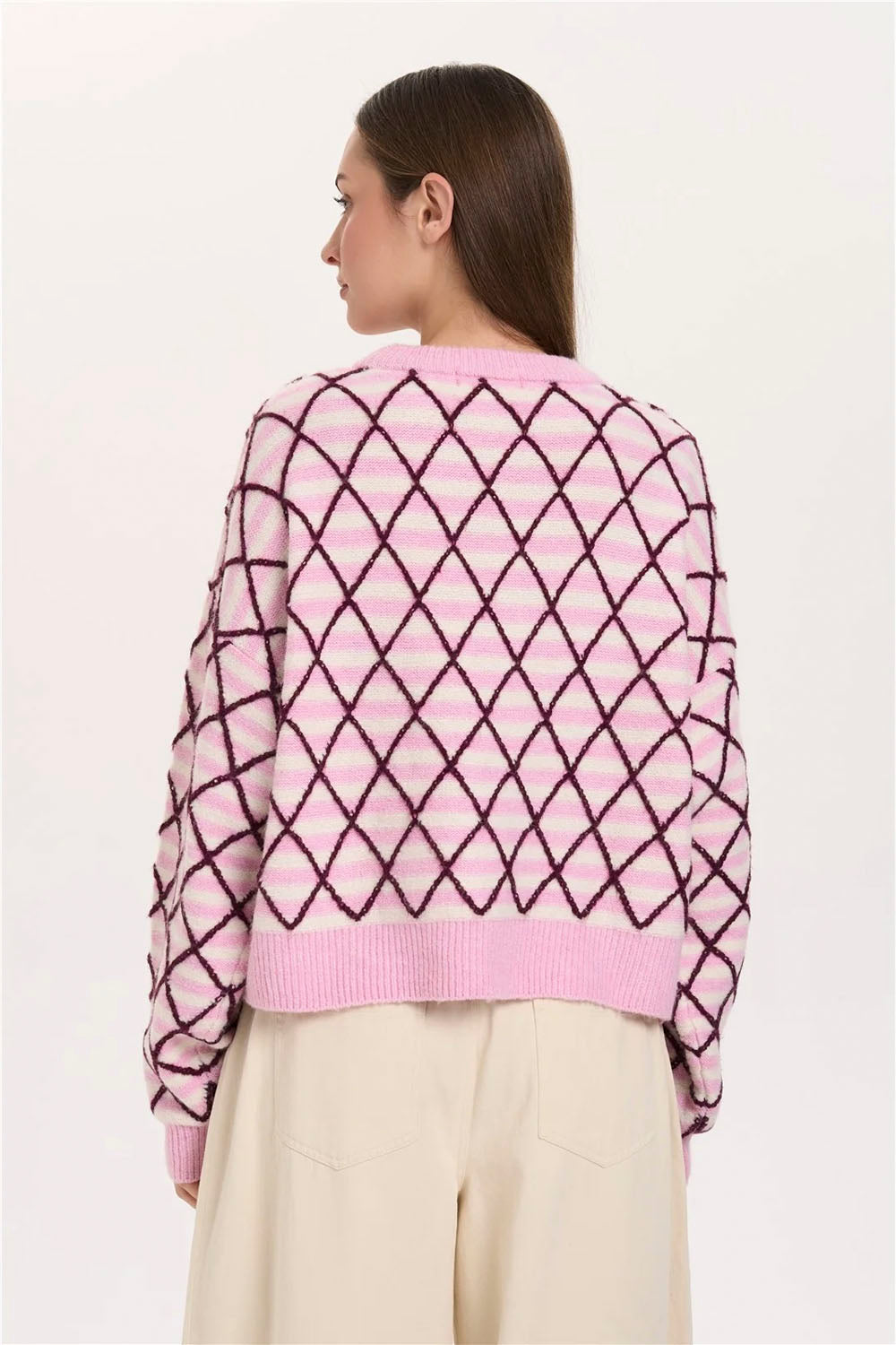 A woman shown from the back wearing a pink knit sweater with a dark purple geometric diamond pattern and cream-colored pants, set against a plain off-white background.